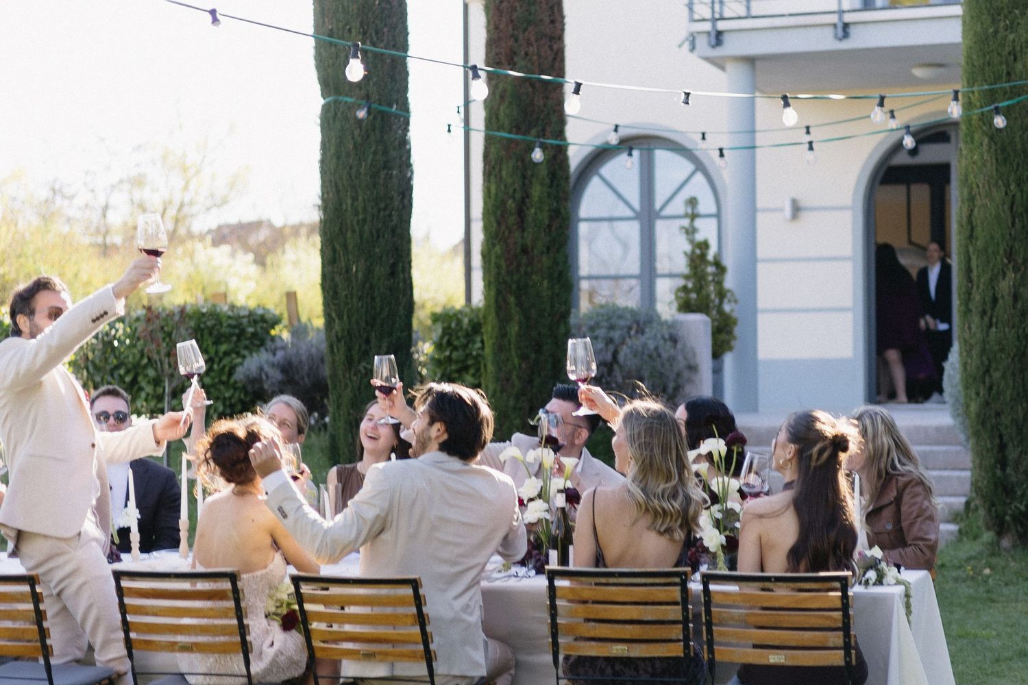 Guests raising glasses in a toast at an elegant outdoor wedding reception with string lights and cypress trees.