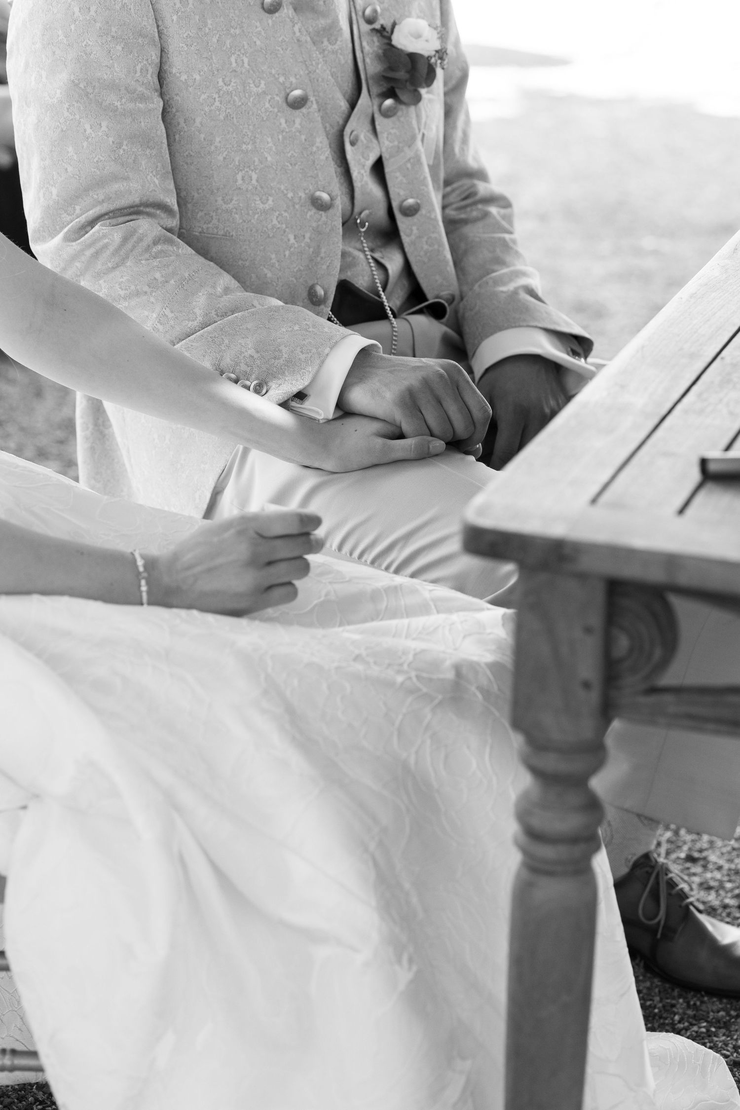Black and white photo of bride and groom holding hands while seated at a wooden table during wedding ceremony.