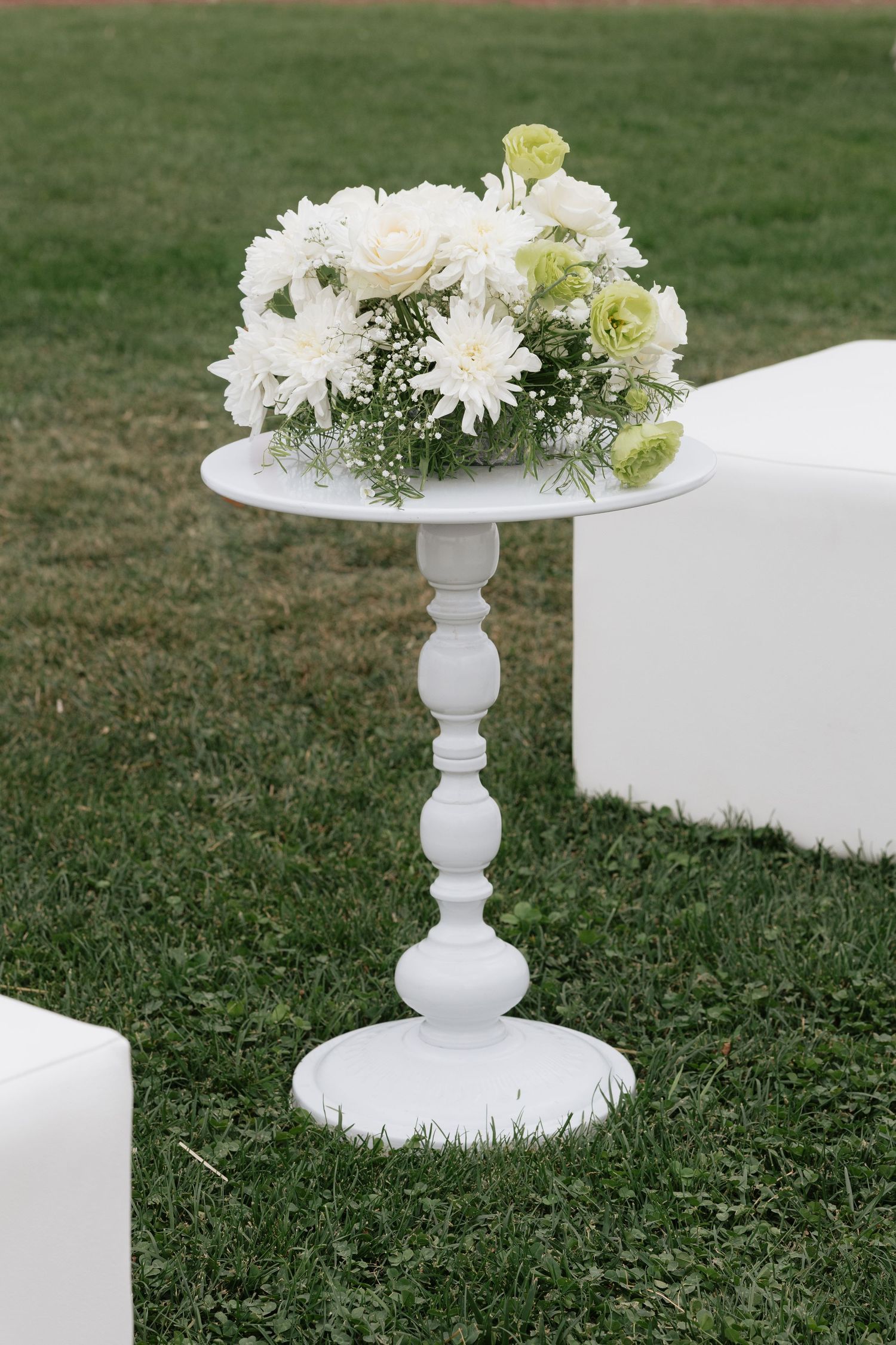 White floral arrangement with roses and chrysanthemums on elegant pedestal table at outdoor wedding ceremony.