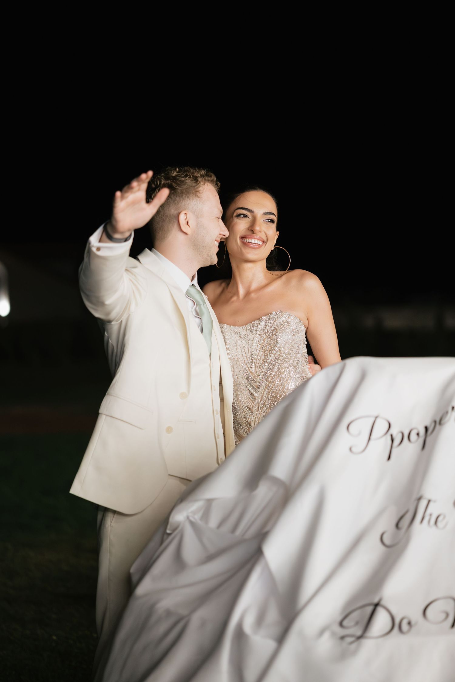 Couple in wedding attire smiling at night, man in cream suit waving, woman in beaded gown holding white fabric.