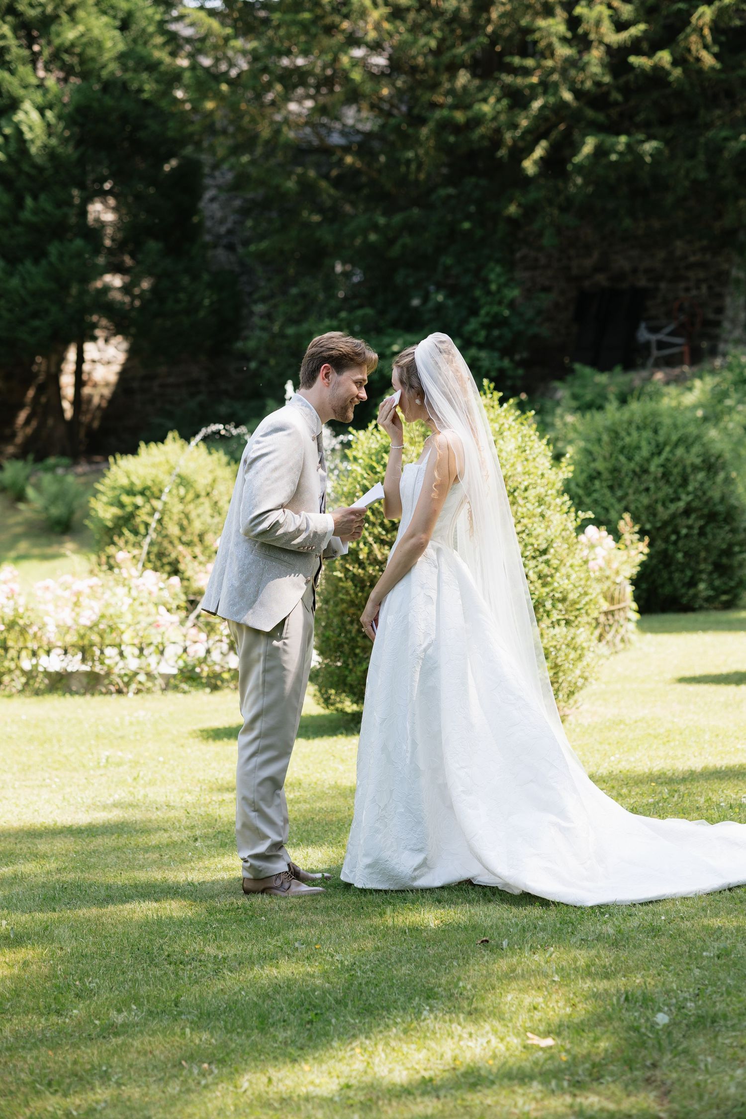 Bride and groom share an intimate moment on a lush green lawn, with the bride in a flowing white gown and veil.