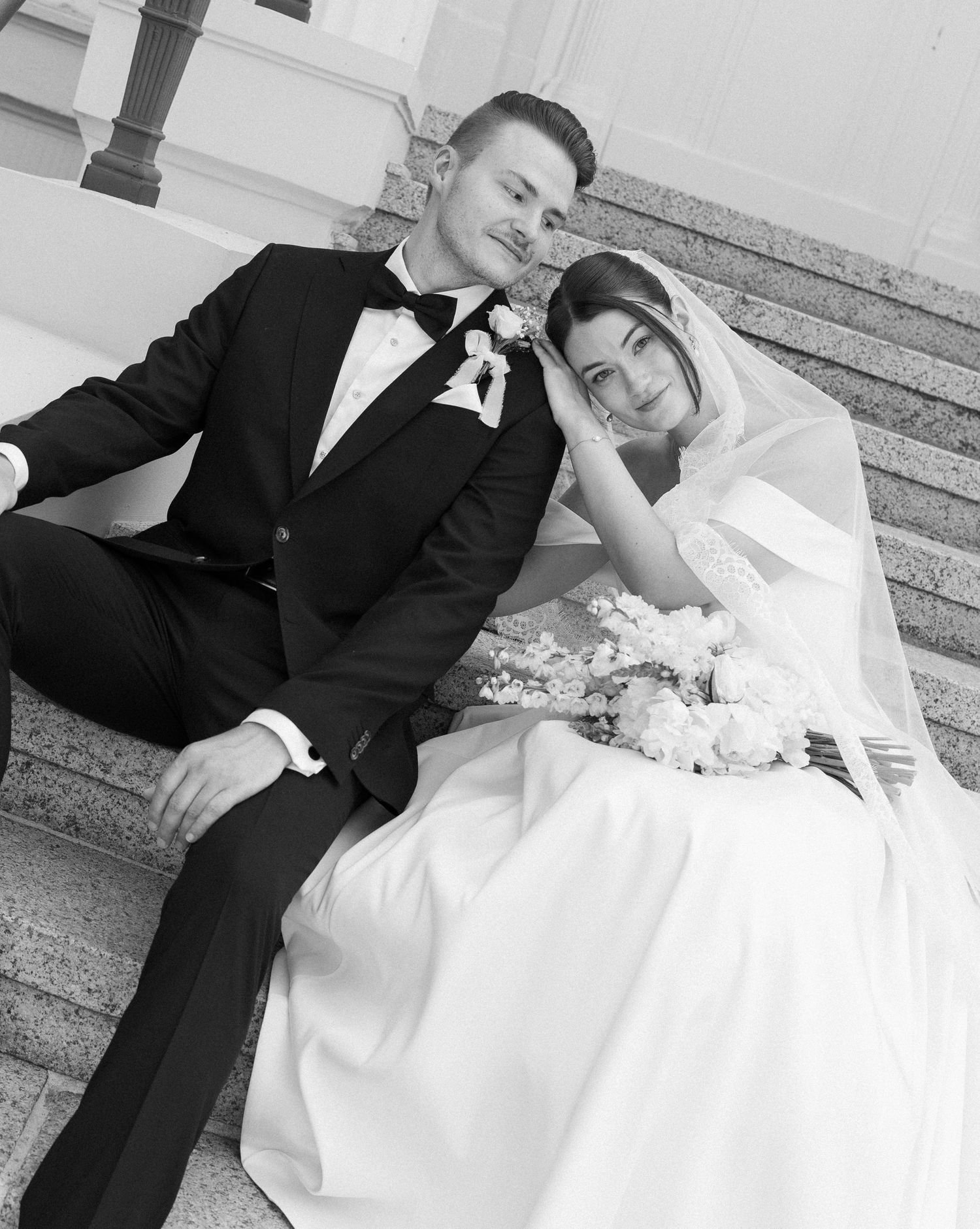Black and white wedding photo of bride and groom sitting on marble steps, bride holding floral bouquet.