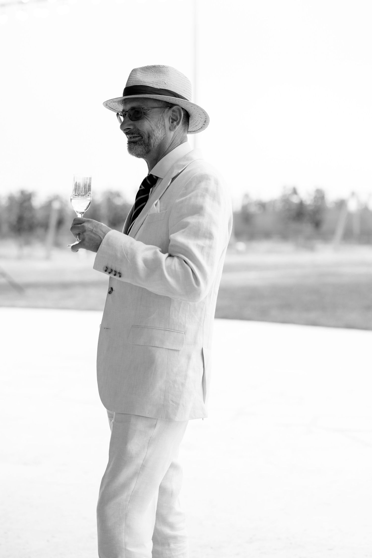 Elegant man in white linen suit and panama hat holding champagne glass outdoors, black and white photo.