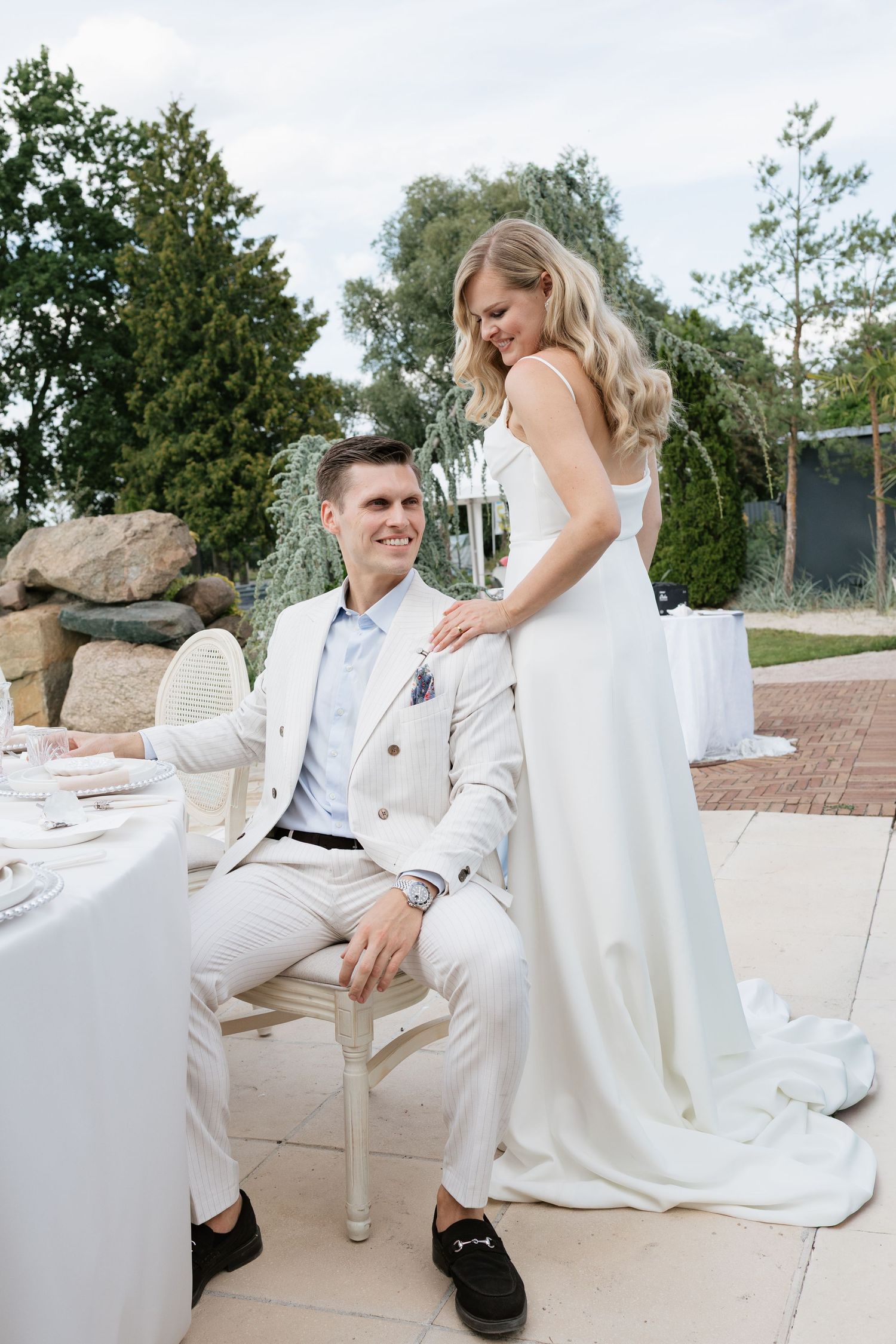 Bride in white gown stands behind smiling groom in cream suit at outdoor wedding reception table.