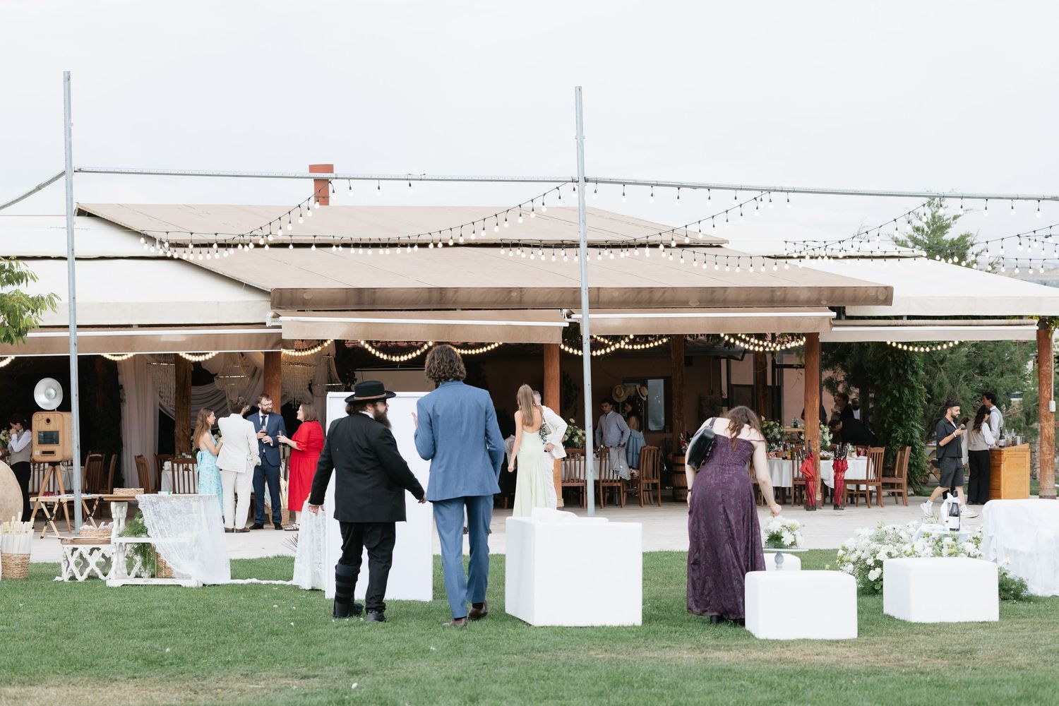 Guests mingle on green lawn at outdoor wedding venue with white decor, string lights, and covered terrace.