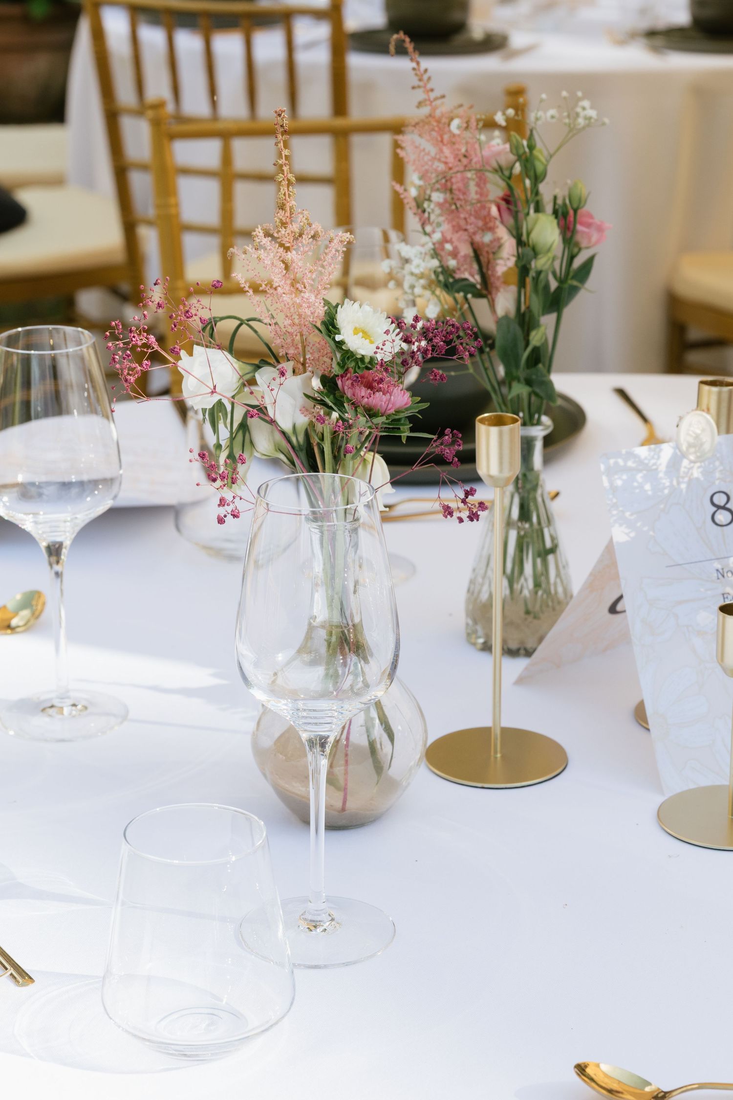 Elegant wedding table setting with gold accents, crystal glasses, and pink floral arrangements in glass vases on white linen.