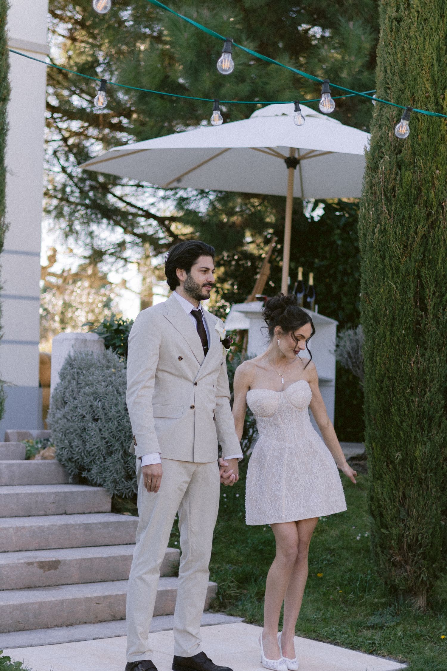 Elegant couple in wedding attire holding hands in a garden, groom in beige suit, bride in short white dress.