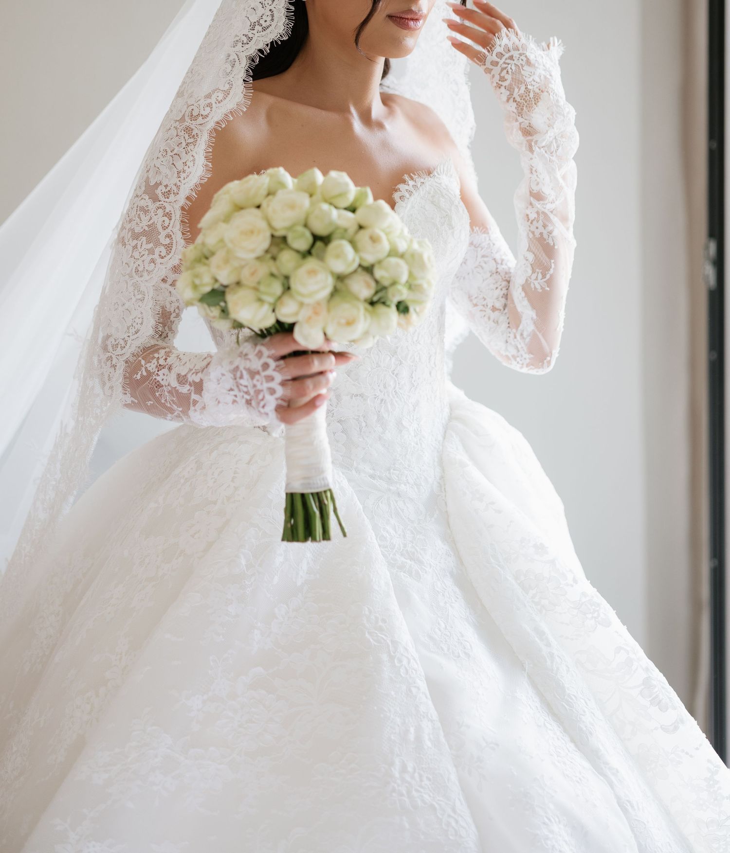 Bride in white lace ball gown with veil holding white peony bouquet near window.