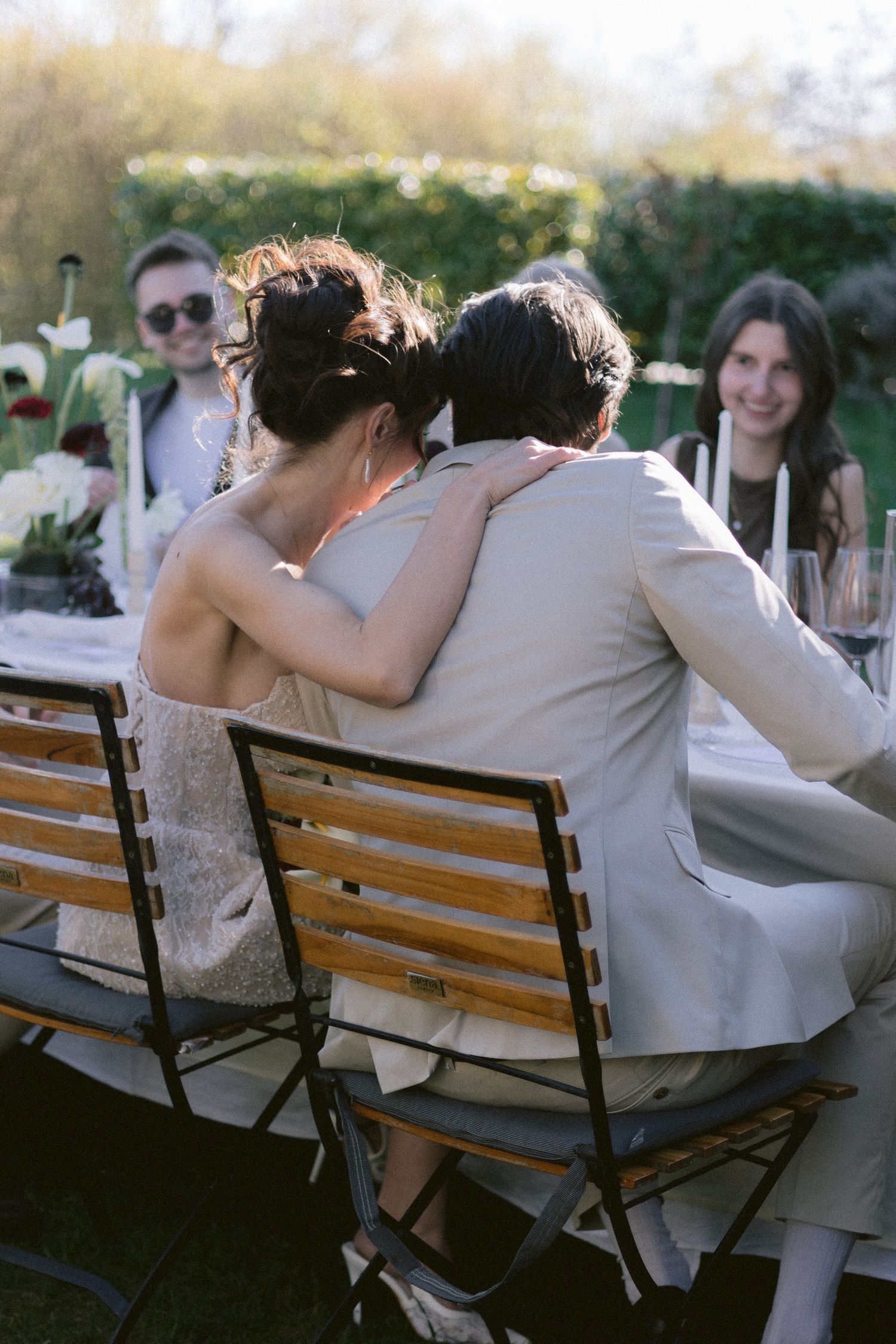 Couple embracing at outdoor wedding reception dinner table with guests smiling in background.