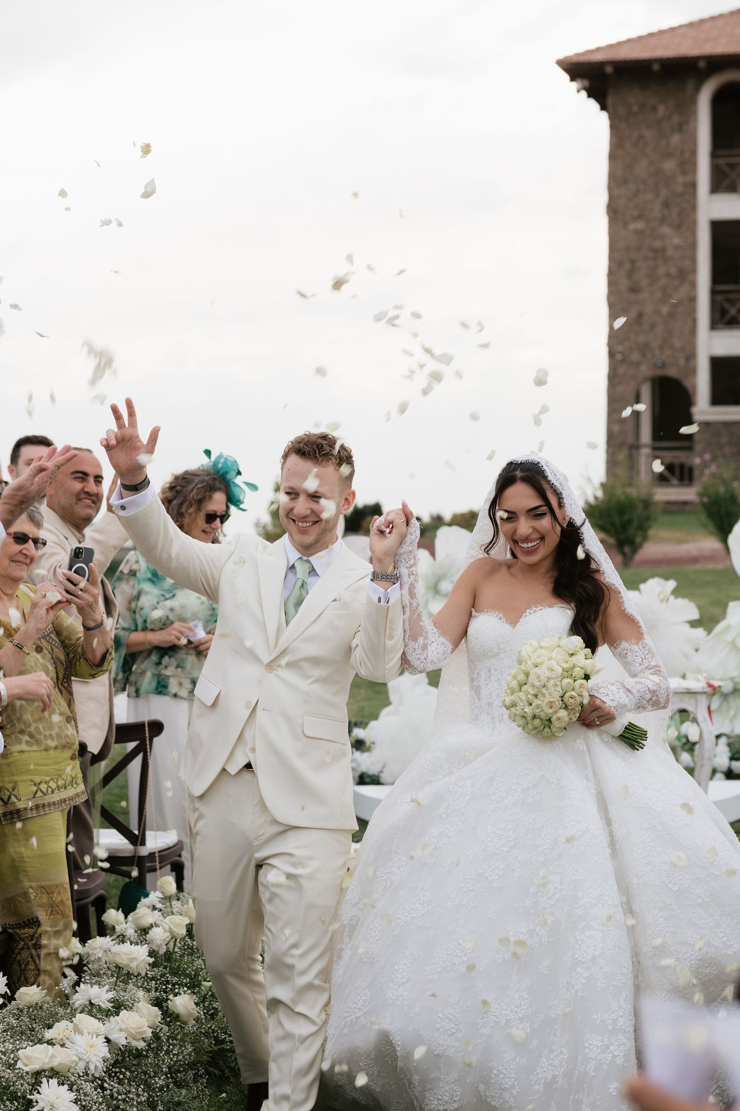 Joyful newlyweds walk down the aisle as guests toss white flower petals at an elegant outdoor wedding ceremony.