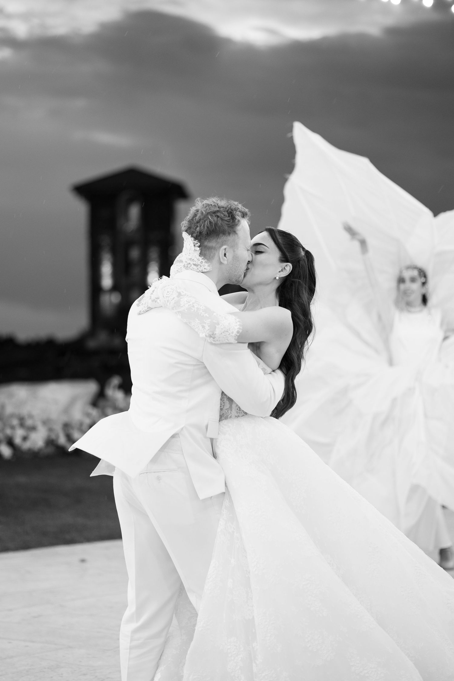 Black and white photo of a couple sharing a kiss at their wedding, bride in flowing gown, groom in white suit.