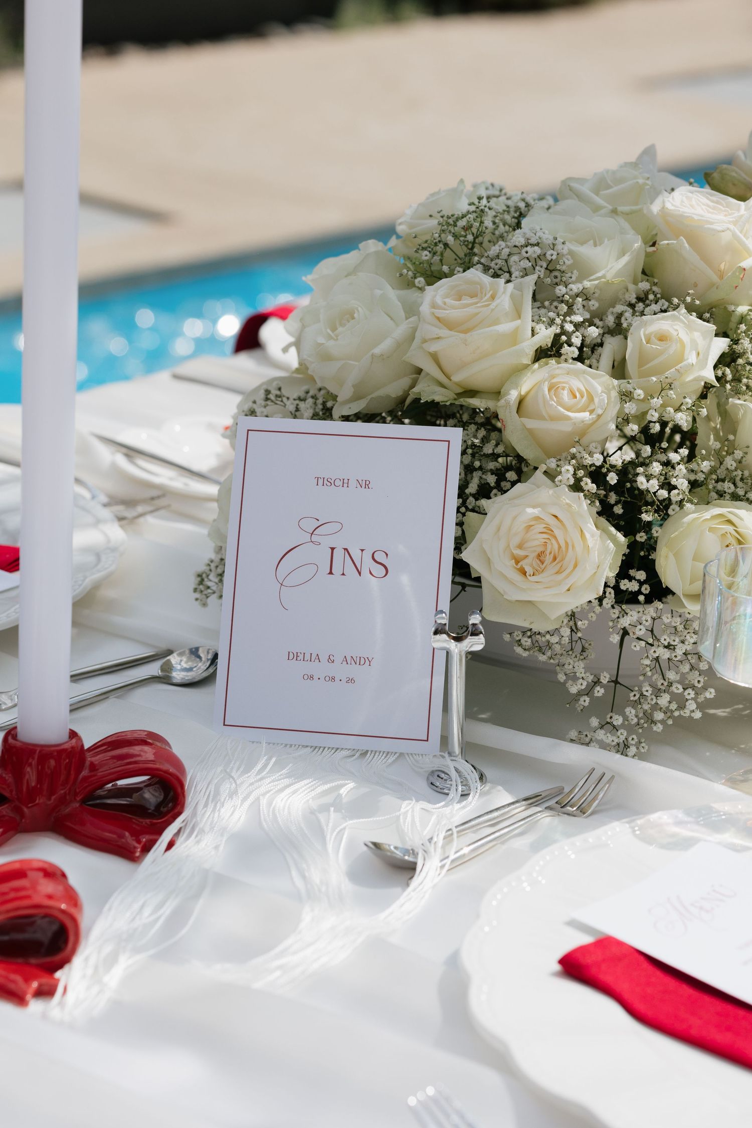 Elegant wedding table with white roses, baby's breath, candles, red napkins, and a table number card reading EINS.