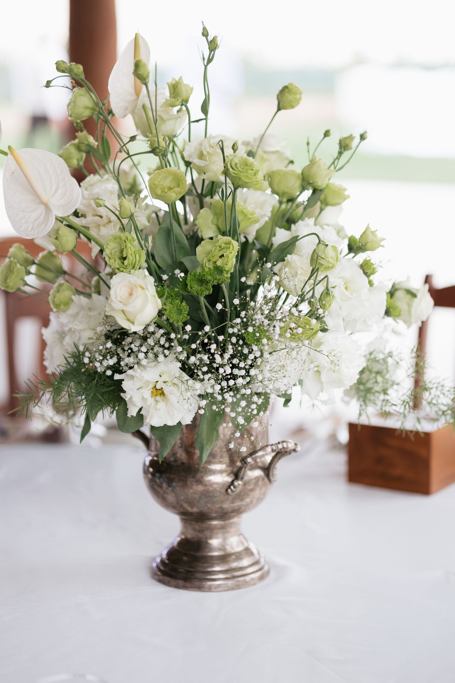 Elegant silver urn vase with white roses, green lisianthus, and baby's breath centerpiece on a white tablecloth.