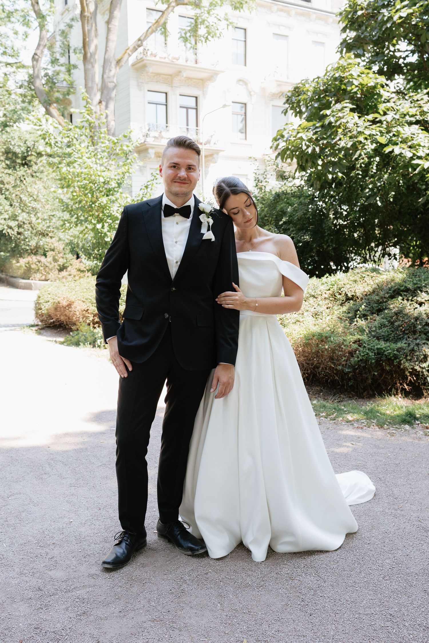 Bride in white off-shoulder gown and groom in black tuxedo pose together in a sunlit garden setting.