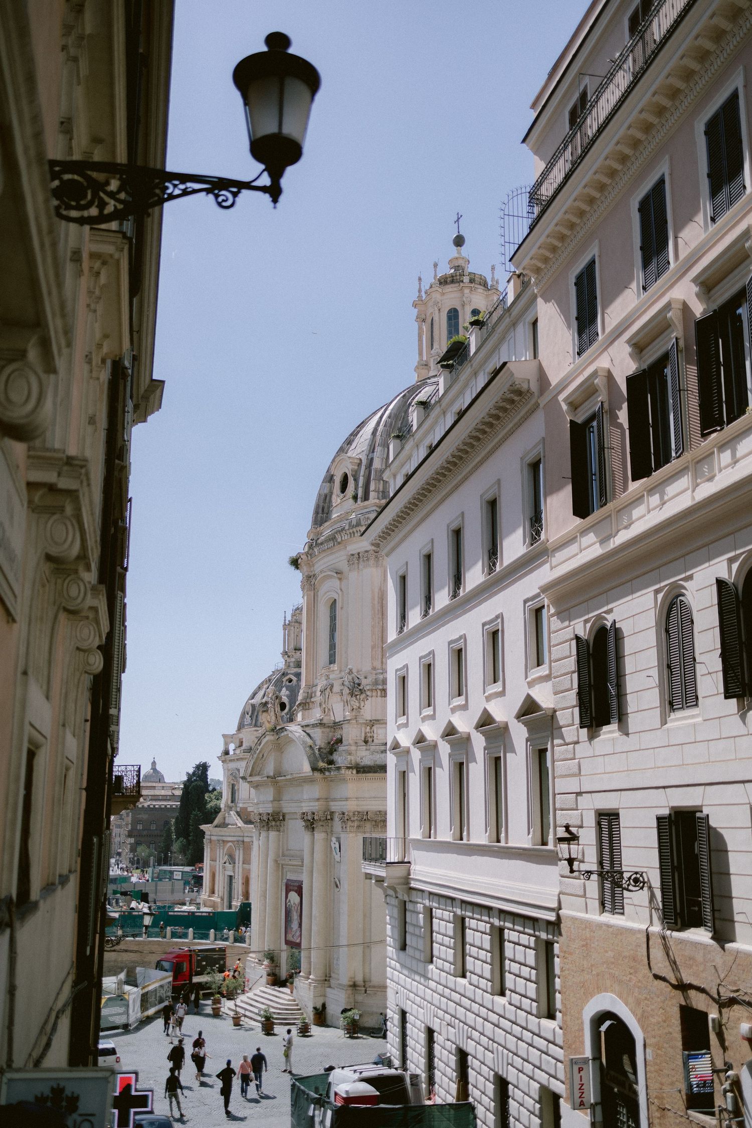 Narrow Roman street with baroque church dome, historic buildings, street lamp, and pedestrians under clear blue sky in Rome, Italy.
