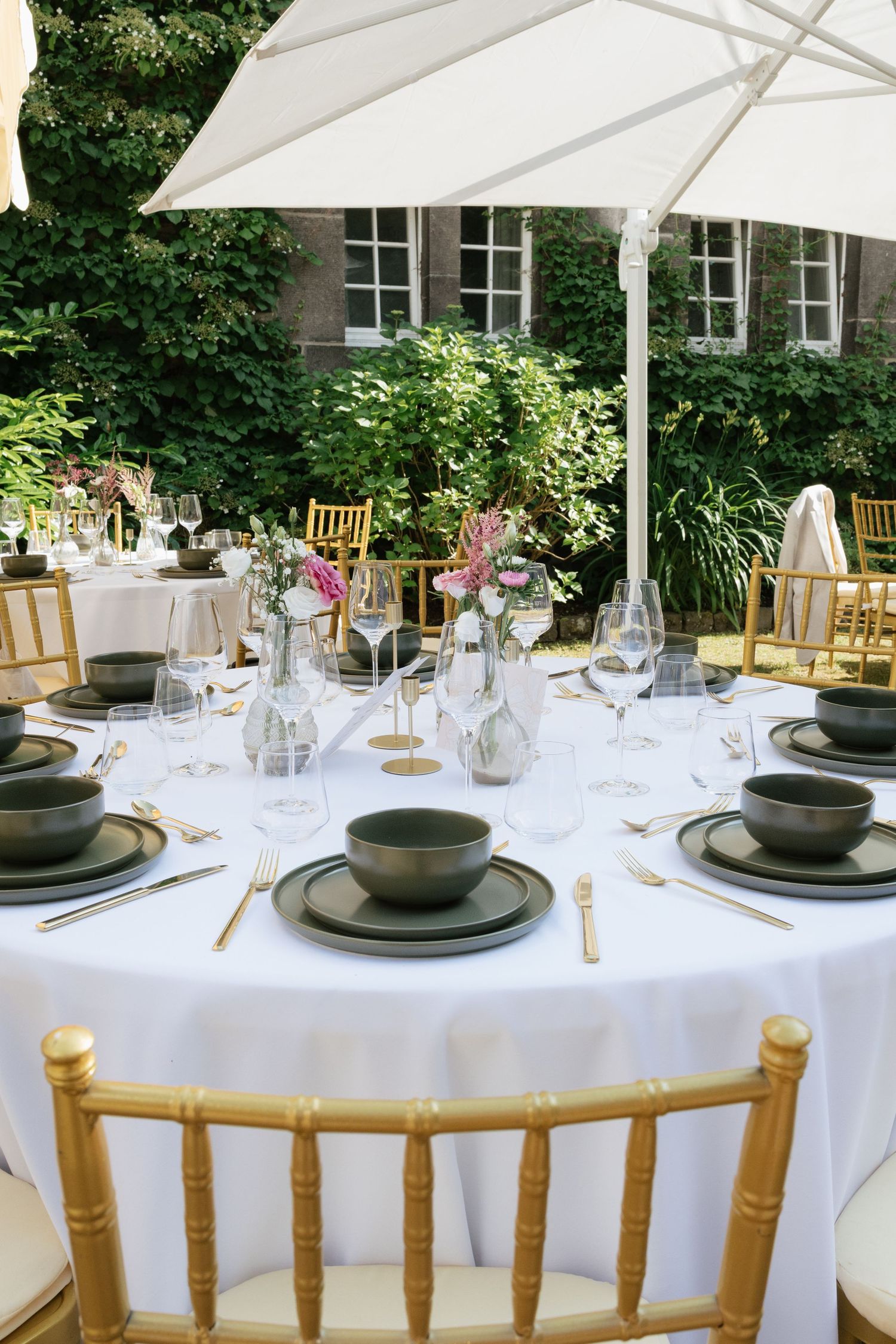 Elegant outdoor wedding reception table set with dark plates, gold cutlery, glassware, and floral centerpieces under a white umbrella.