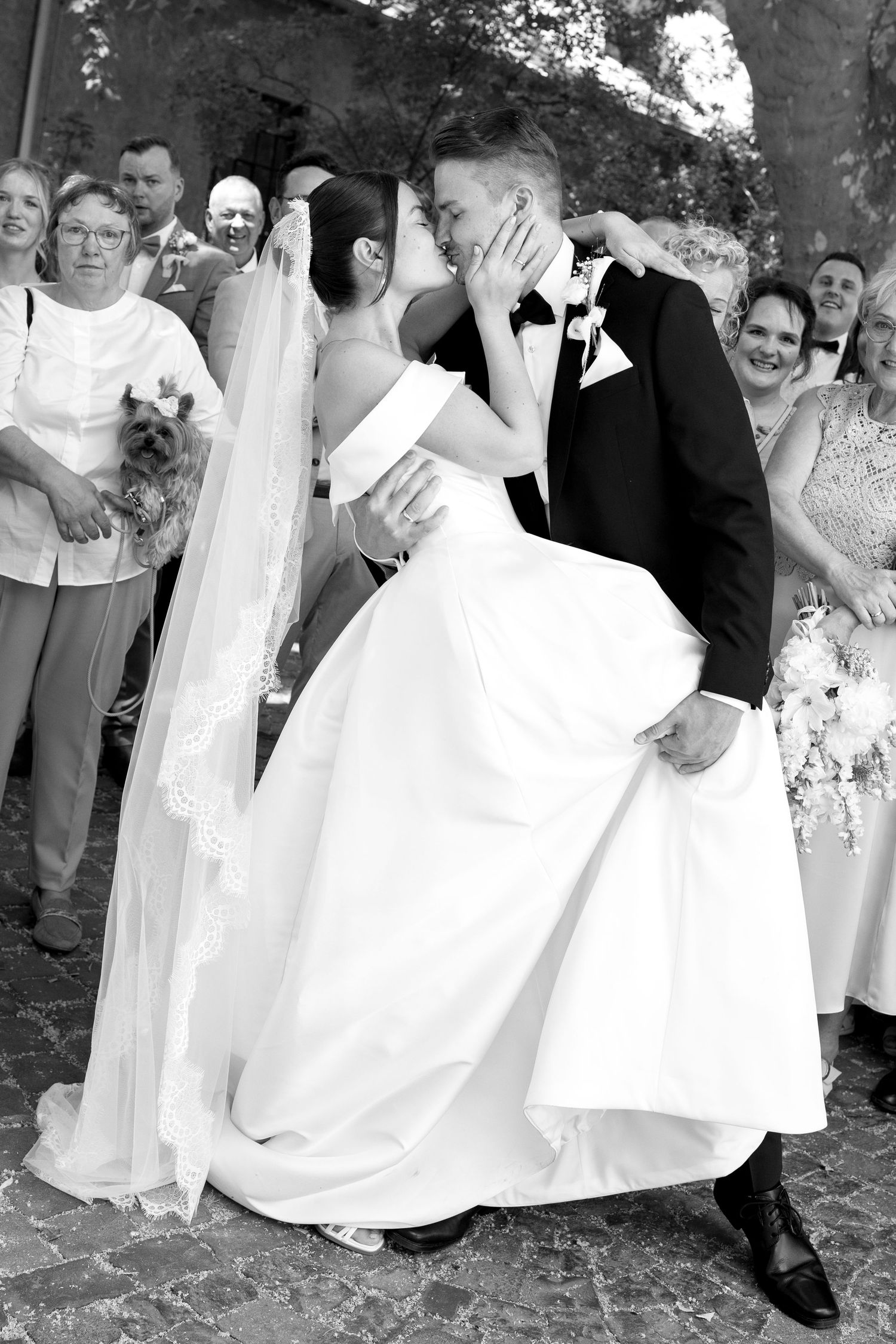 Black and white photo of a groom lifting his bride in a white gown and veil, sharing a kiss surrounded by wedding guests.