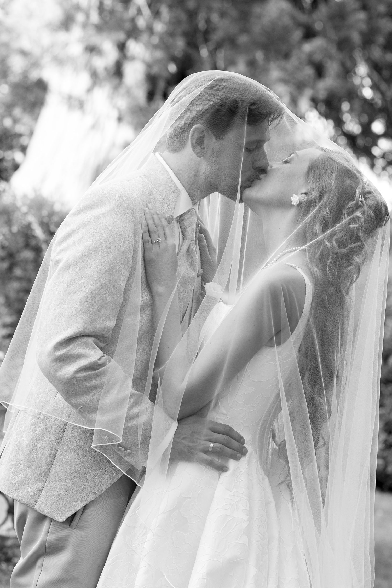 Black and white photo of a bride and groom sharing a kiss under a flowing veil outdoors on their wedding day.