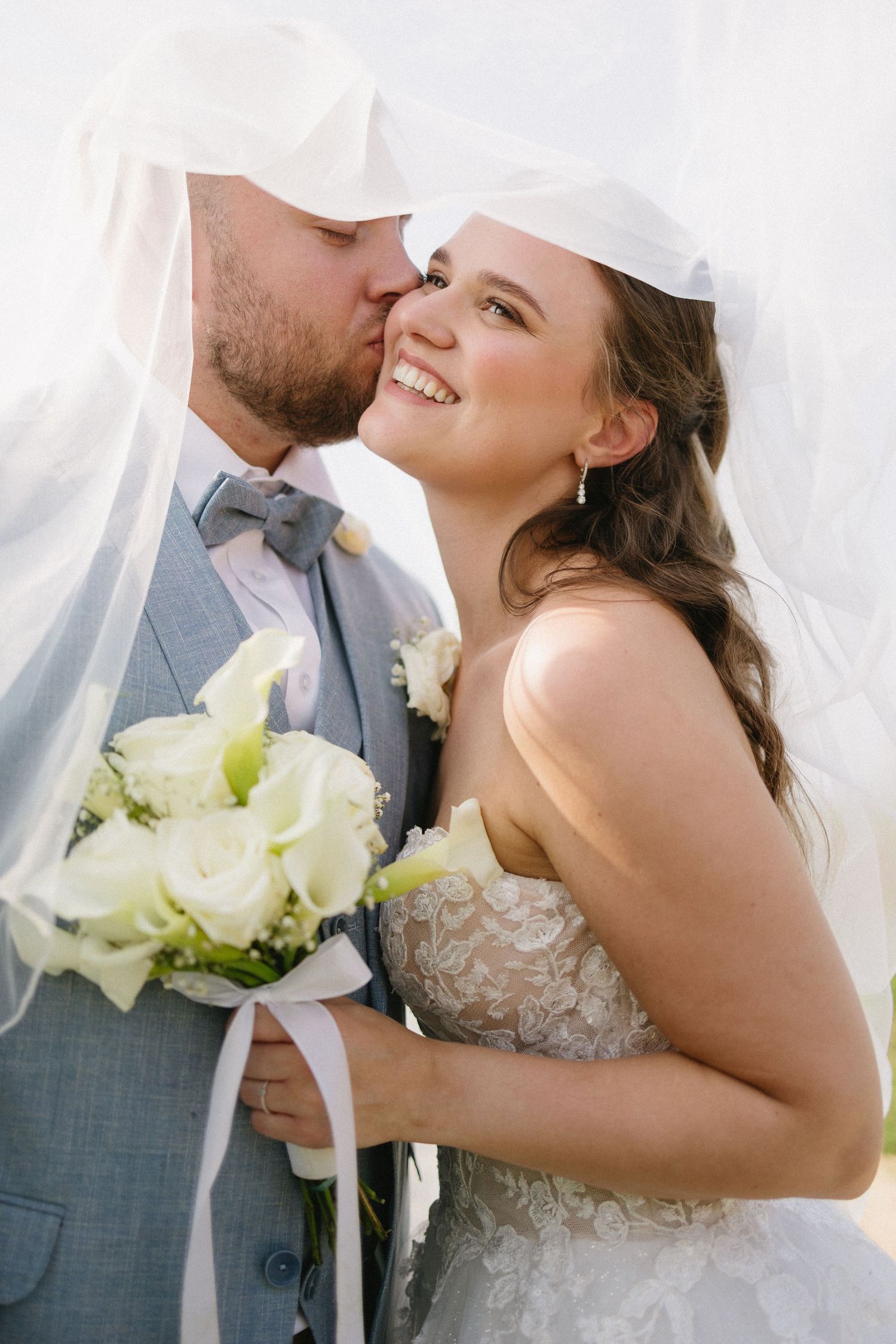 Groom kissing bride on cheek under veil, bride smiling holding white rose bouquet in lace wedding dress.