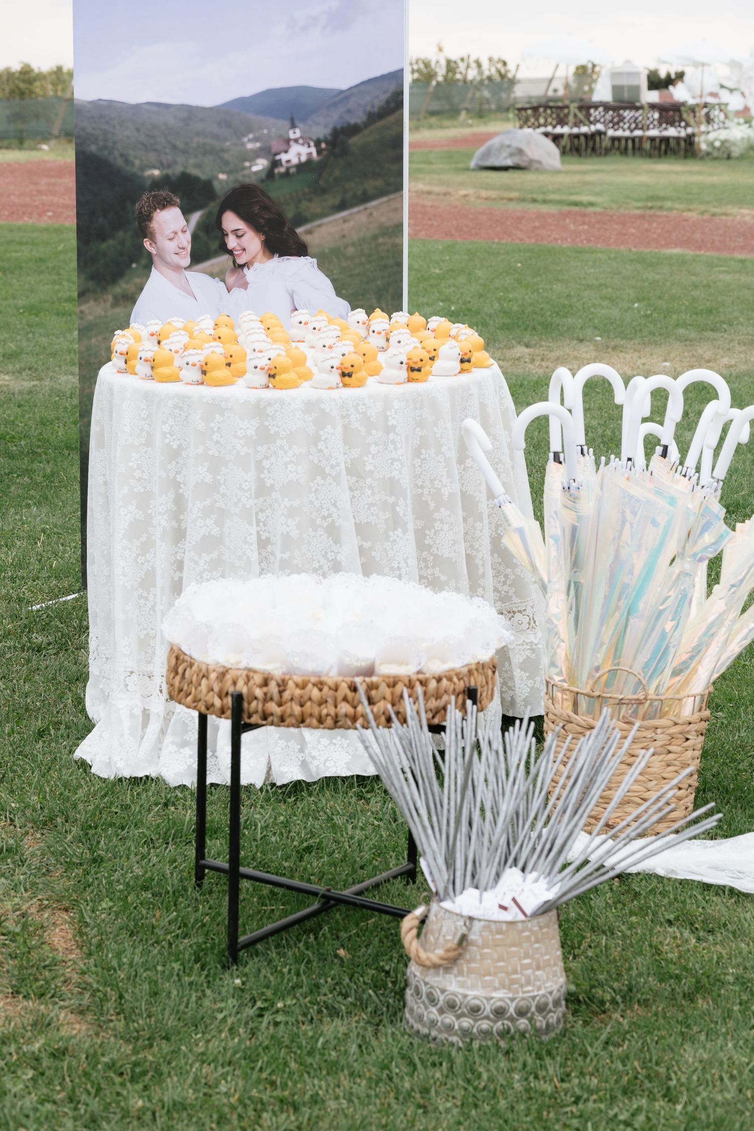 Outdoor wedding display with rubber ducks on lace table, couple photo backdrop, and white umbrellas in wicker basket on grass.