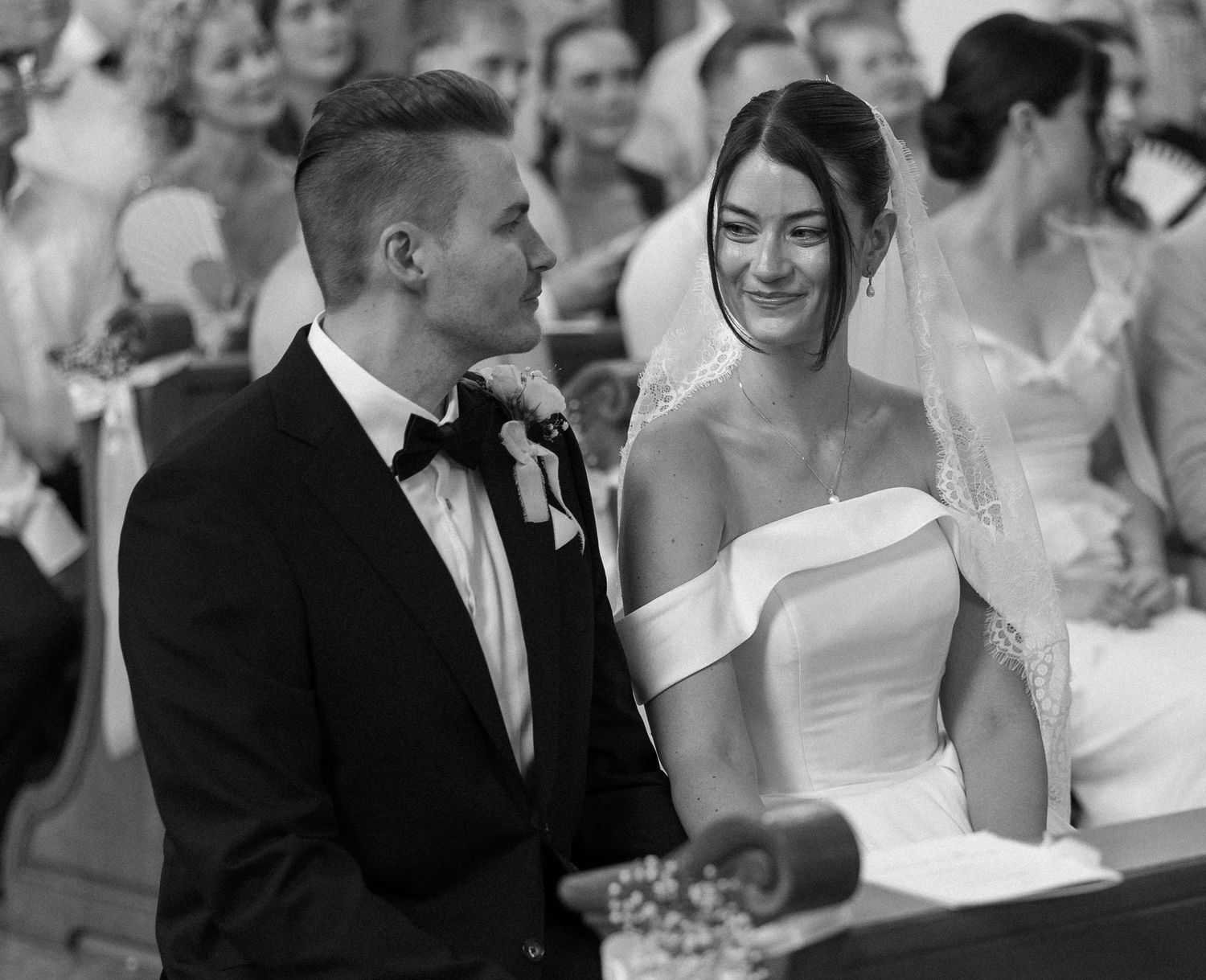 Black and white wedding photo of a smiling bride in veil and groom in tuxedo standing at the altar.