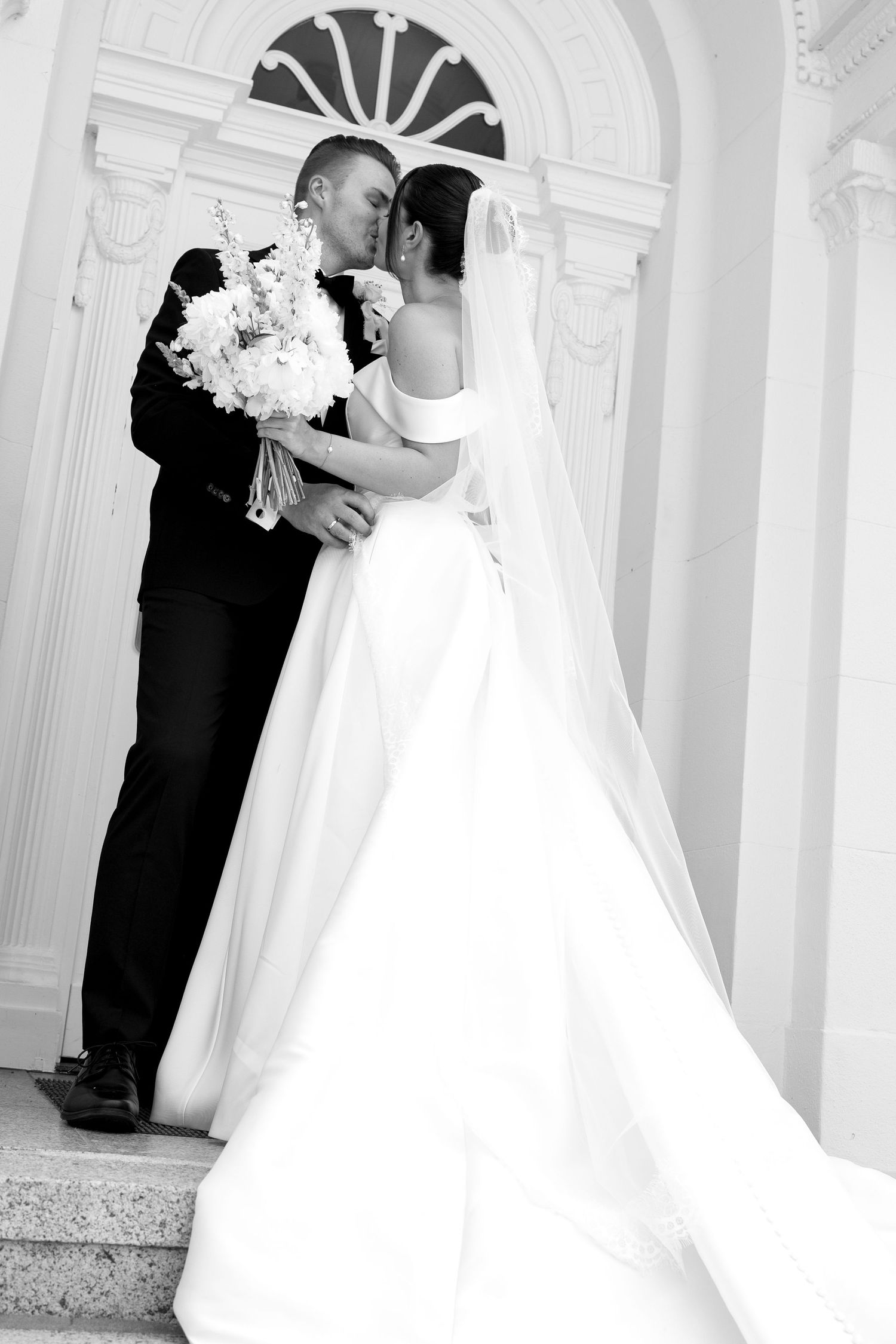 Black and white photo of a bride and groom sharing a kiss on church steps, bride holding a bouquet.