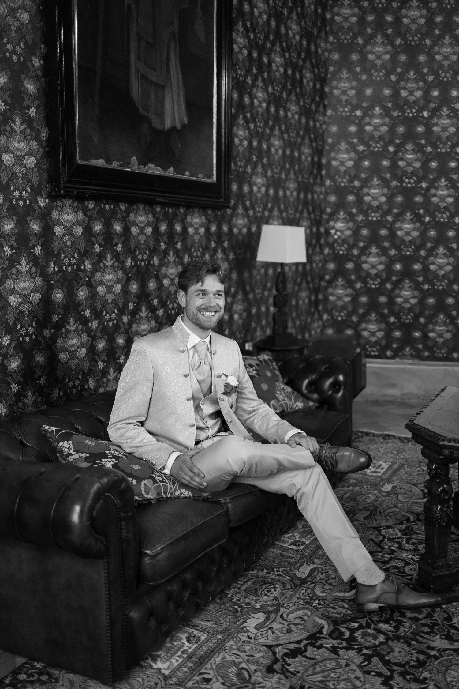 Smiling groom in light suit sits on leather sofa in elegant room with patterned wallpaper, black and white photo.