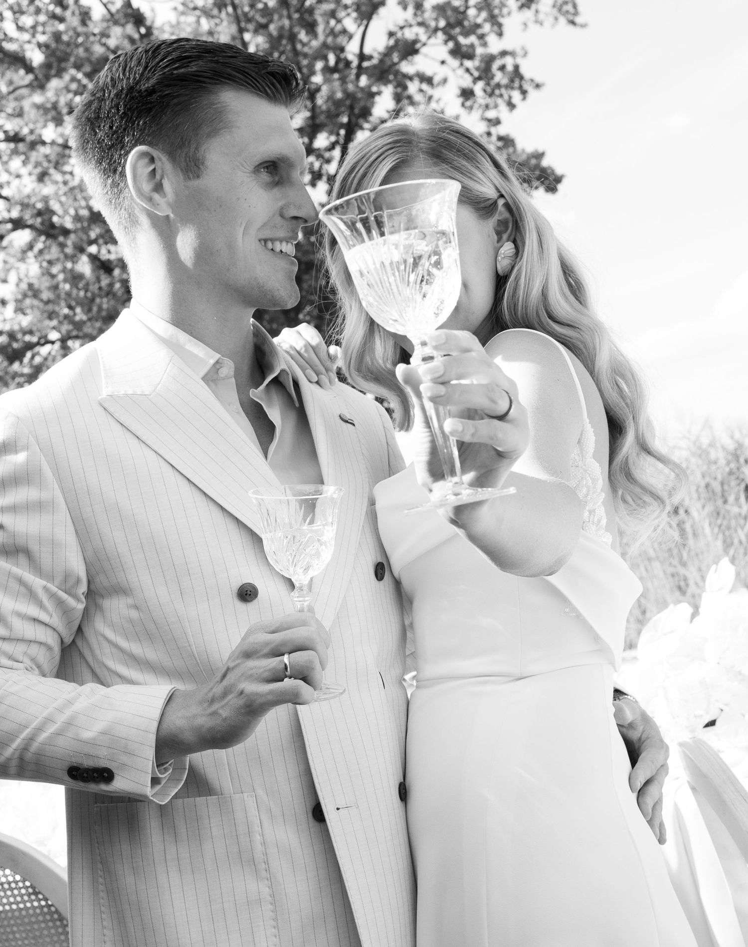 Black and white photo of a couple in elegant attire toasting with crystal glasses at an outdoor celebration.