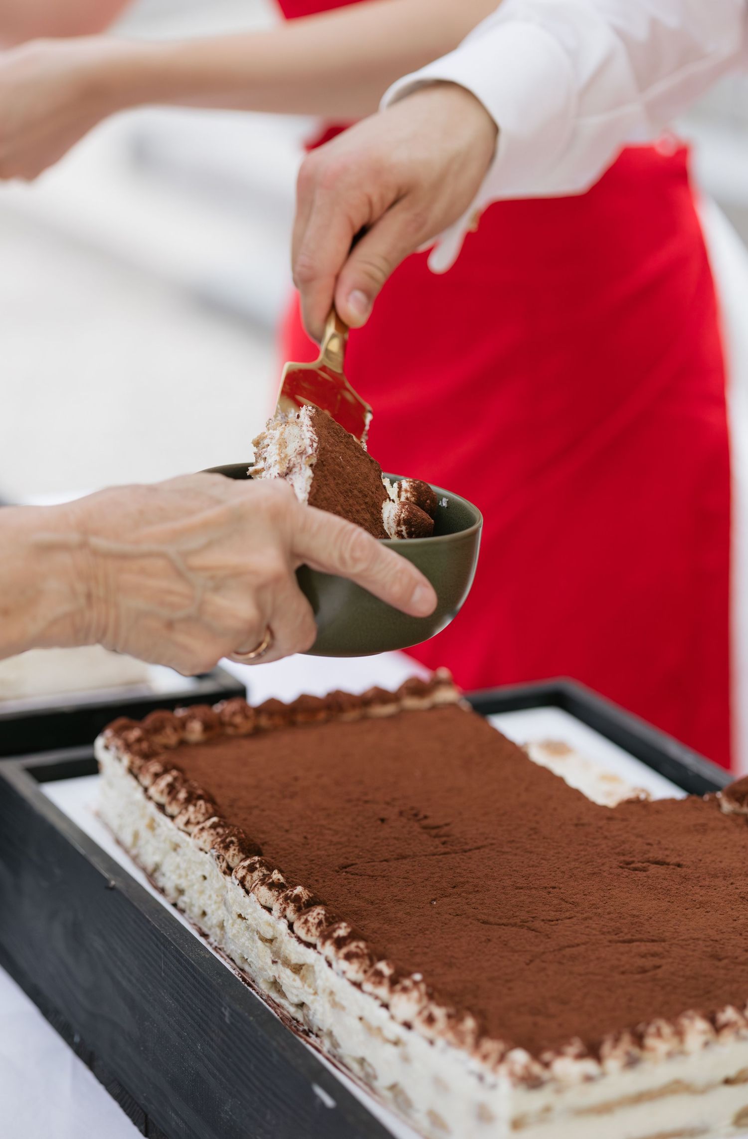 Hands serving a slice of tiramisu cake dusted with cocoa powder into a small bowl at a catering event.
