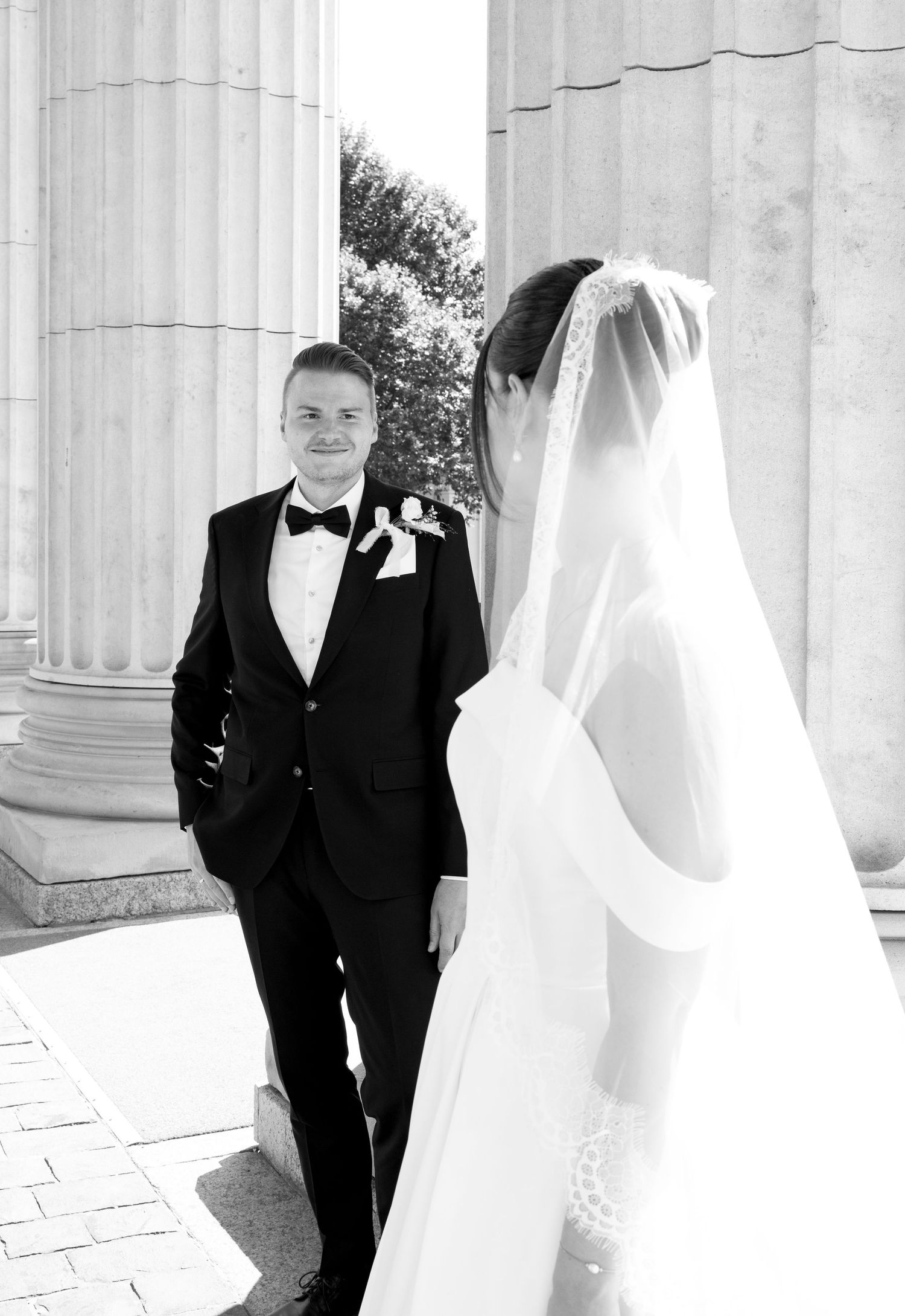 Black and white wedding photo of groom in tuxedo smiling at bride in veil beside grand columns.
