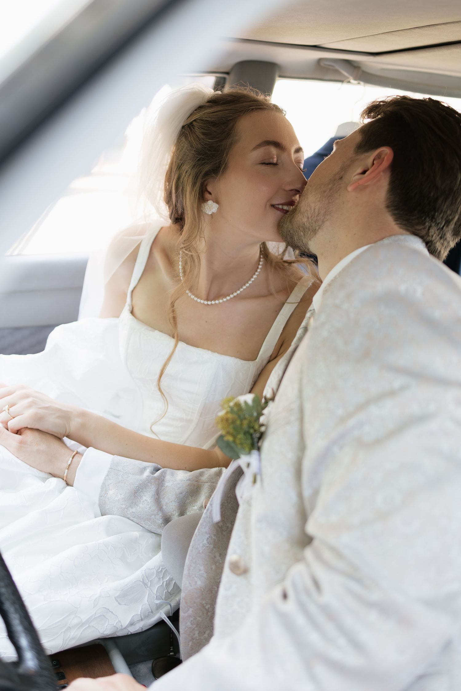 Newlywed bride and groom sharing a romantic kiss inside a car, bride wearing white dress and veil with pearl necklace.