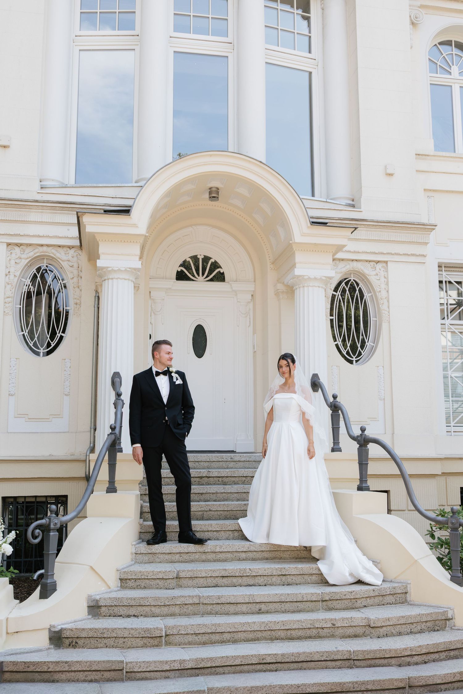 Bride and groom pose on grand white mansion steps, groom in tuxedo, bride in elegant off-shoulder ball gown.