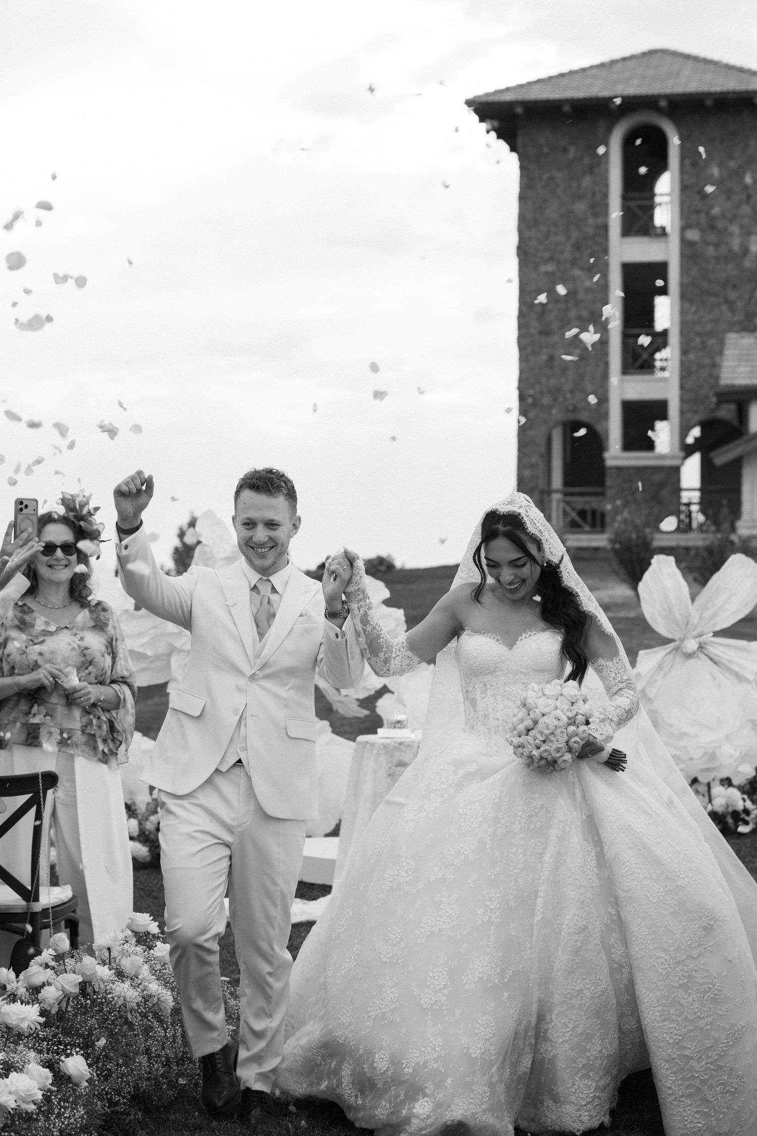 Black and white photo of a joyful bride and groom walking down the aisle as guests toss confetti at their outdoor wedding.