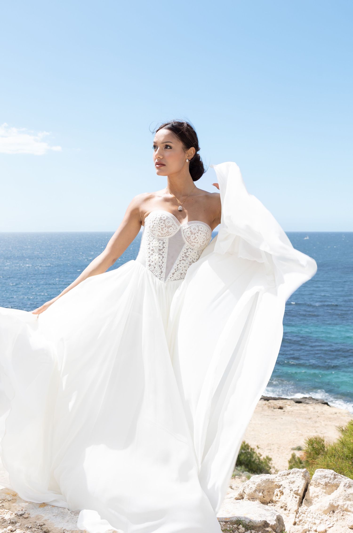 Bride in flowing white strapless wedding gown with lace bodice, standing on rocky cliff with turquoise ocean backdrop.