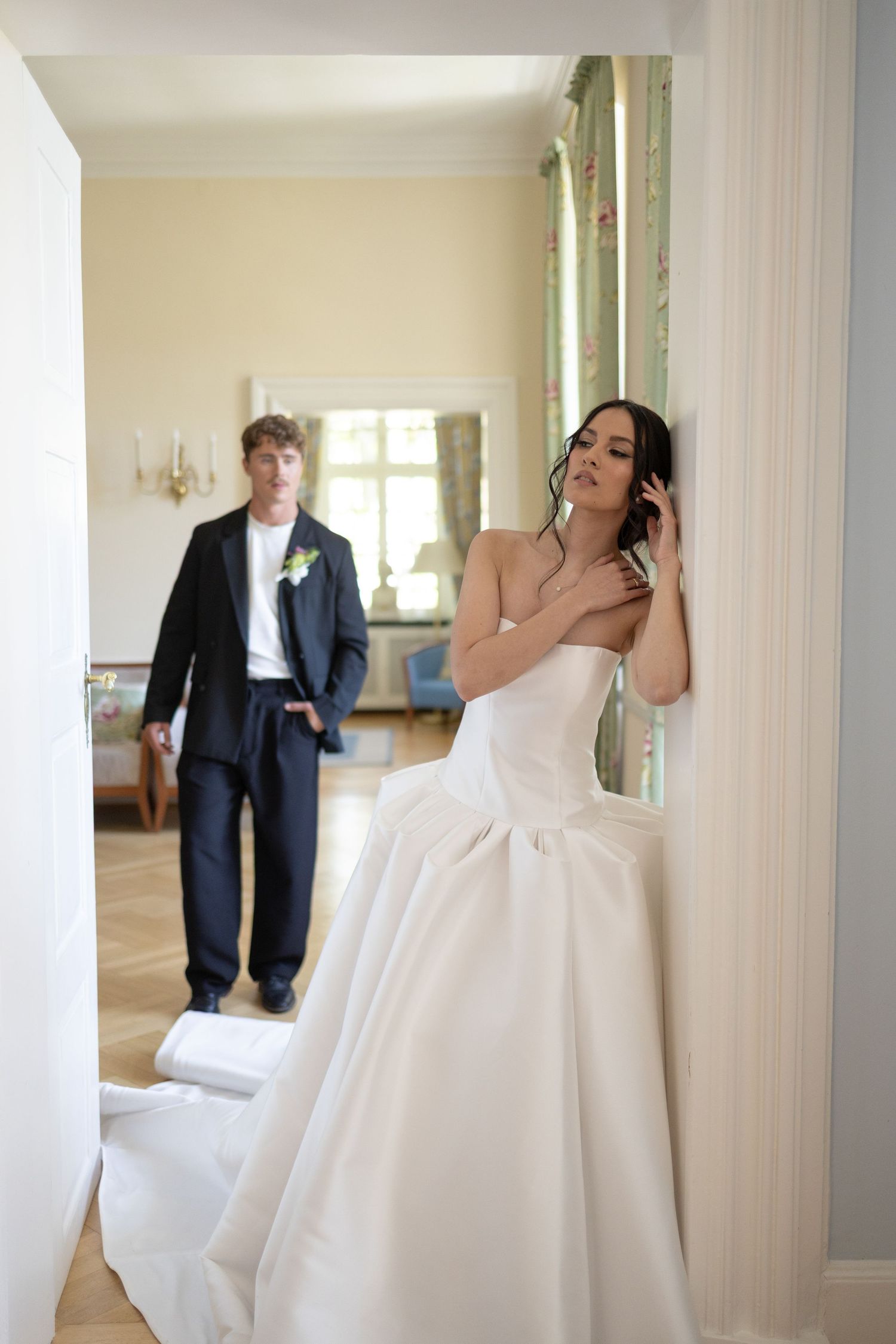 Bride in strapless white gown leans against pillar as groom in navy suit stands in elegant room.