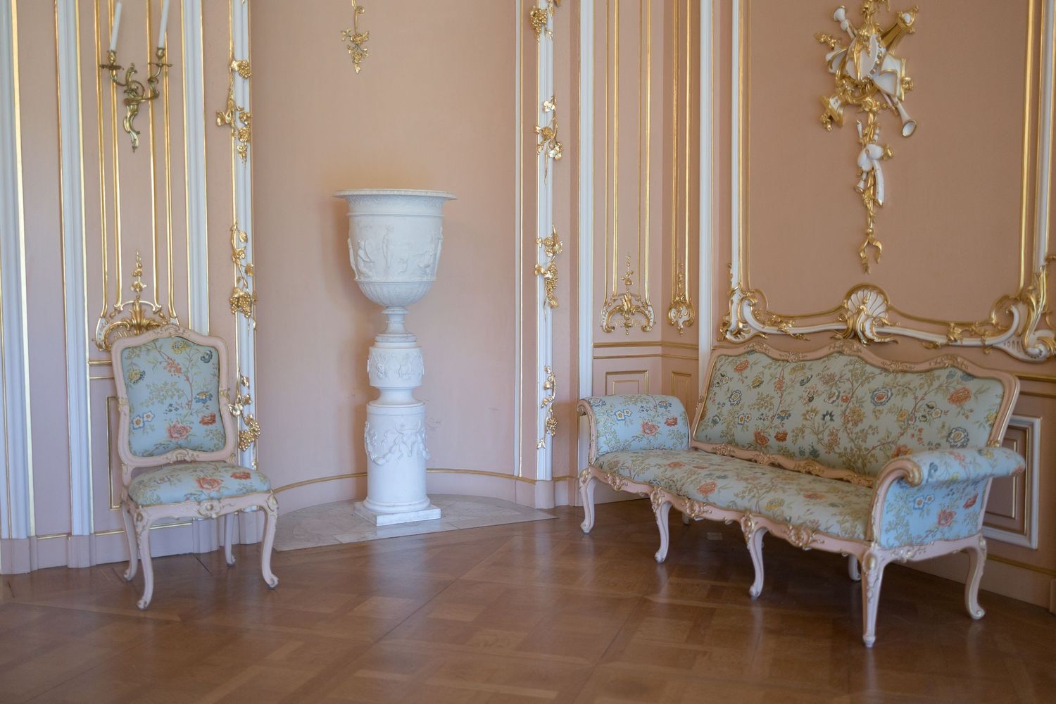 Ornate Rococo room with gilded moldings, floral upholstered settee, chair, and white urn pedestal on parquet floor in Kassel Wilhelmsthal..