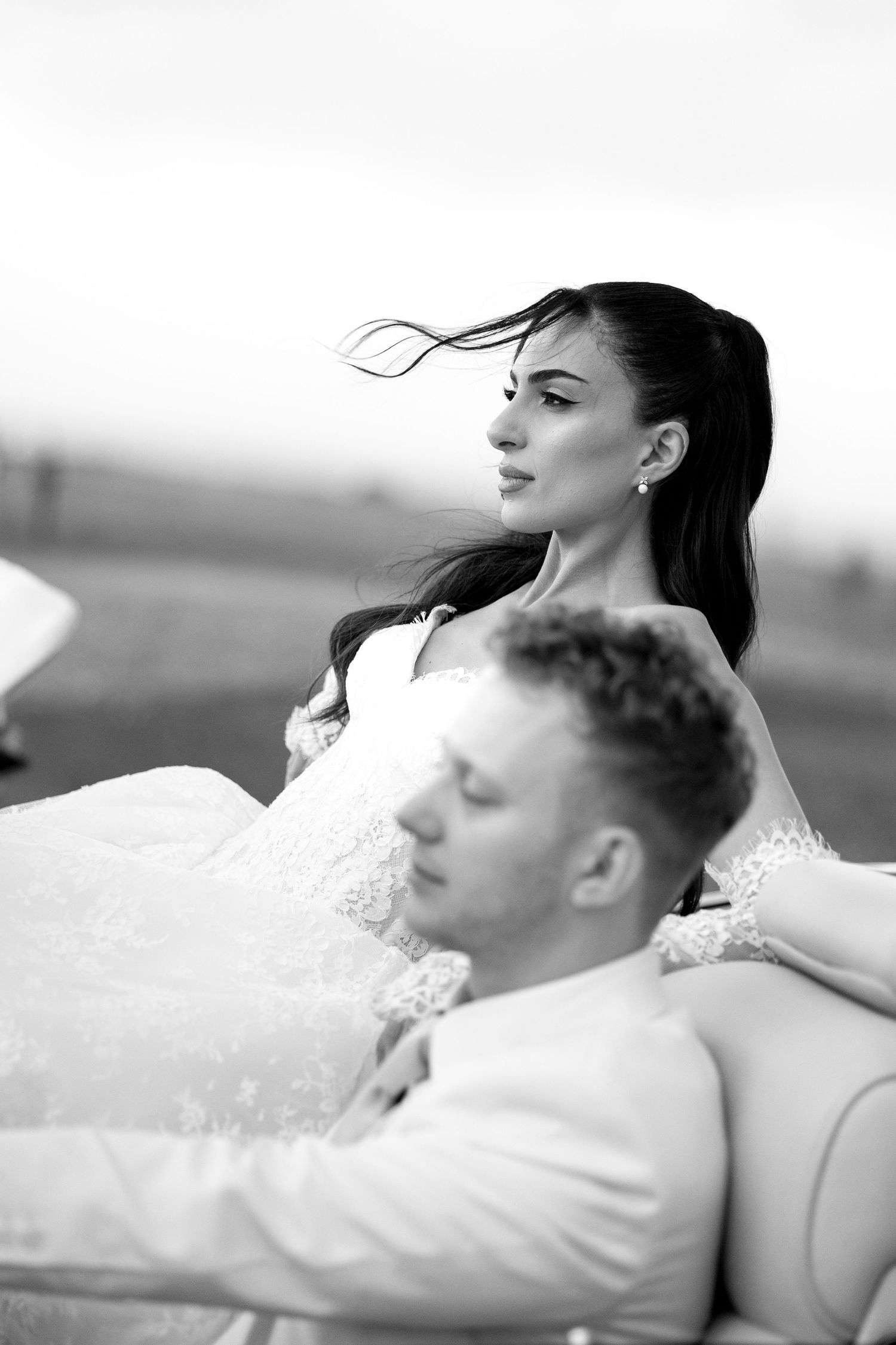 Black and white photo of a bride and groom sitting together outdoors, her hair blowing in the wind.