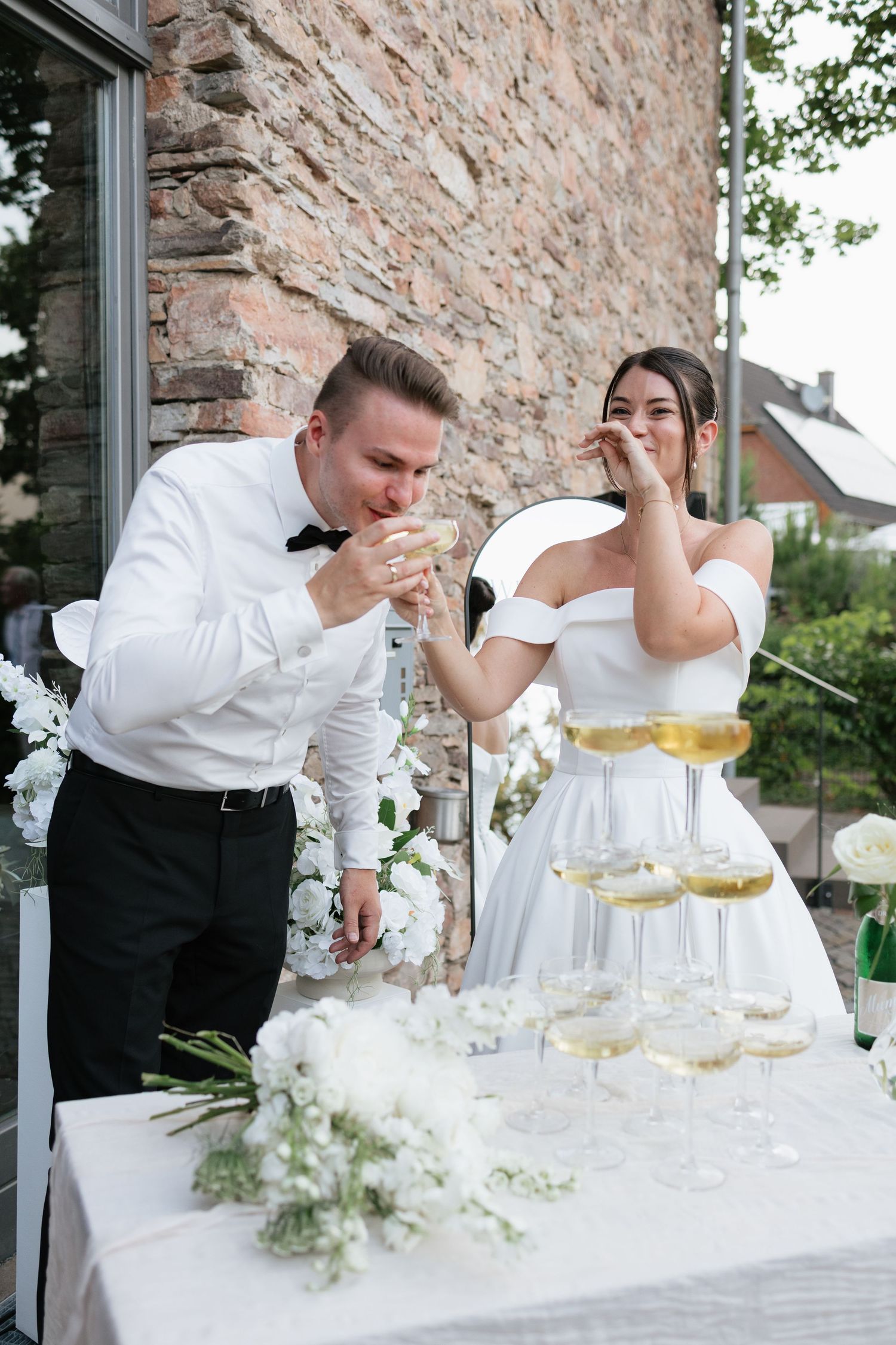 Bride and groom sipping champagne at wedding reception beside elegant champagne tower with white floral arrangements.