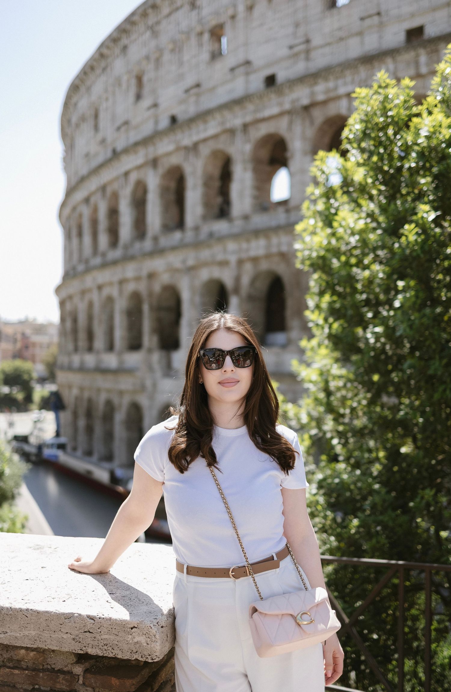 Woman in white outfit with crossbody bag posing in front of the Colosseum in Rome on a sunny day.