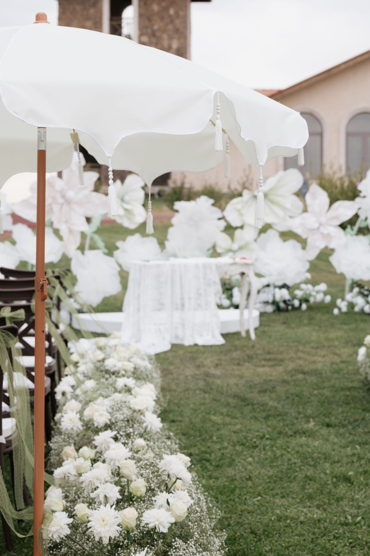 Elegant outdoor wedding aisle lined with white roses and baby's breath, white umbrellas, and large floral decorations on green lawn.