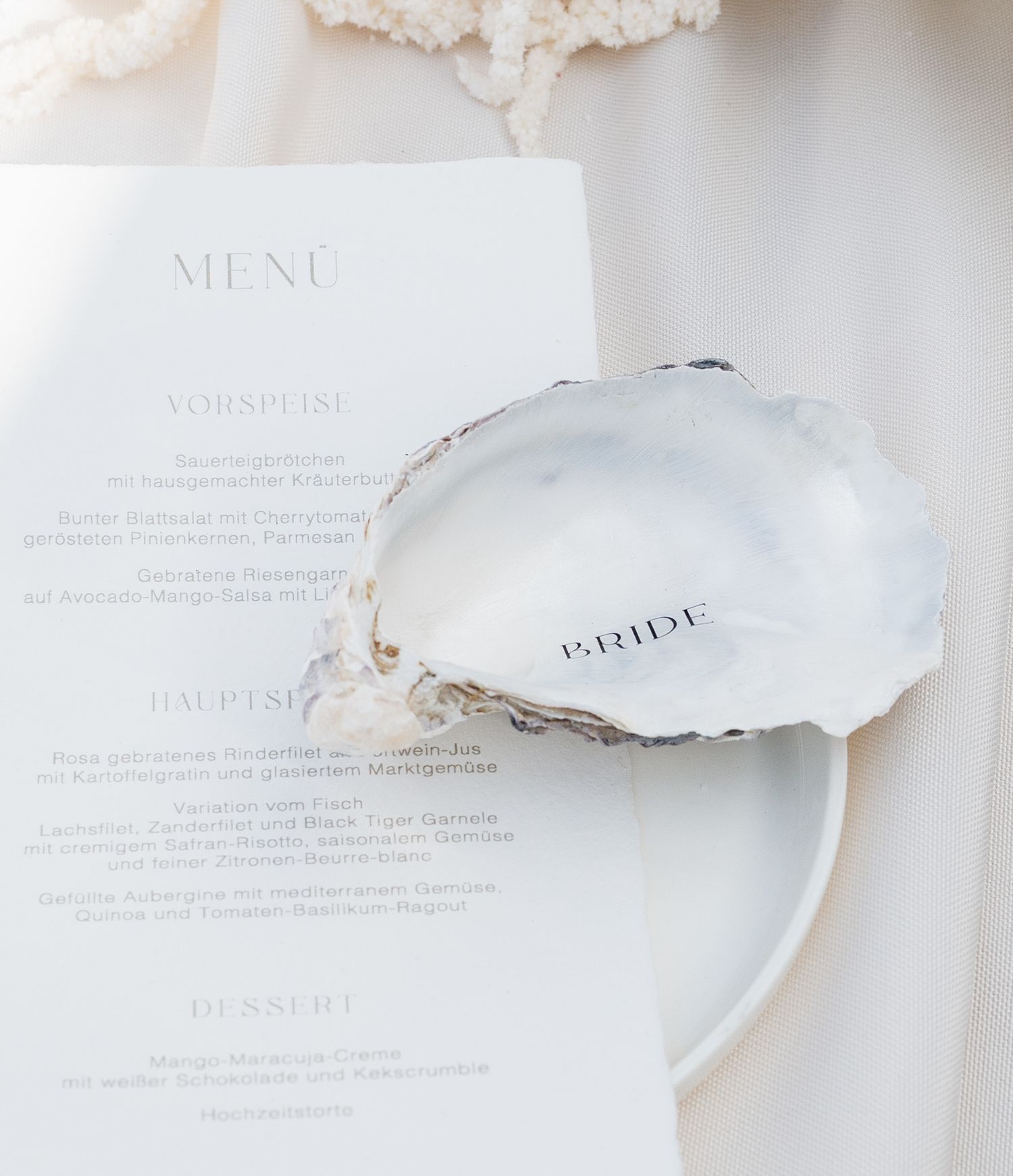 Oyster shell place card with 'Bride' text resting on a white wedding menu card, elegant bridal table setting.