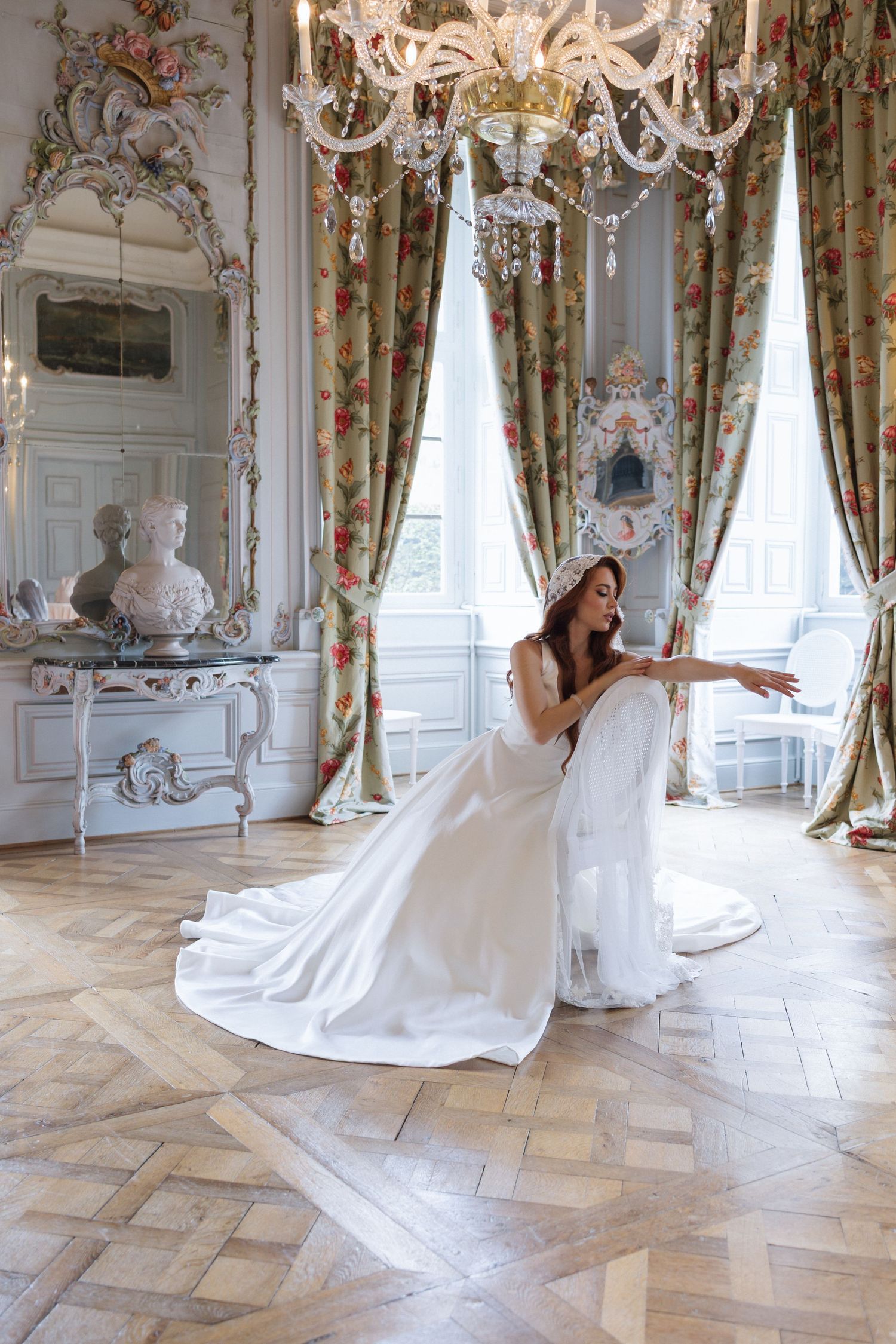 Bride in white gown poses gracefully in ornate French chateau room with crystal chandelier and floral curtains.
