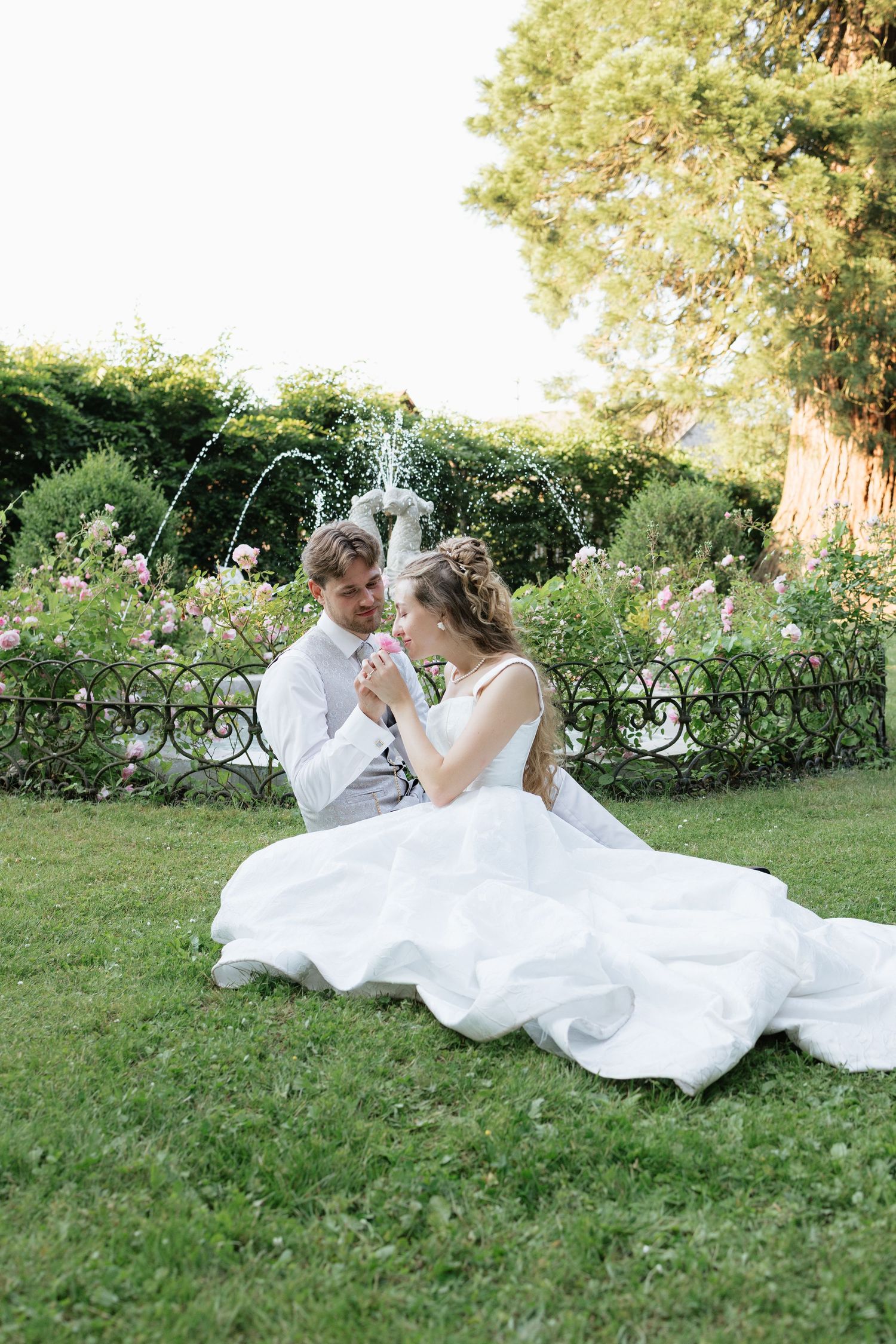 Bride and groom sitting on green grass in front of a fountain and rose garden on their wedding day.