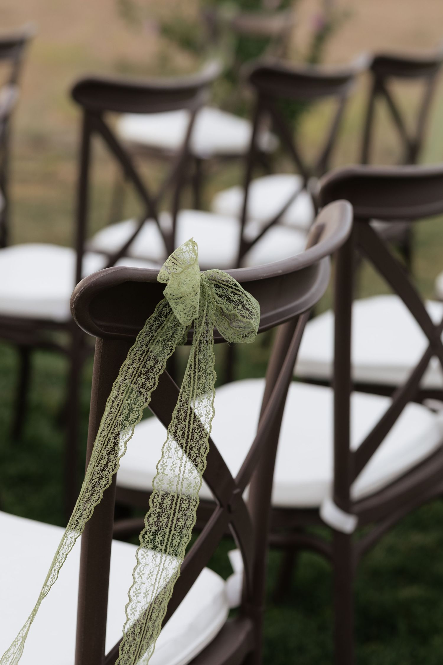 Dark brown cross-back wedding chairs with elegant green lace ribbon bows arranged outdoors for a garden ceremony.