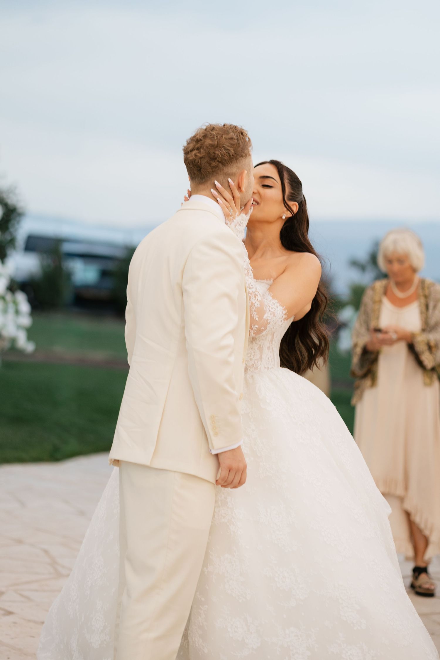 Bride and groom share first kiss in elegant white wedding attire outdoors at dusk with guests in background.
