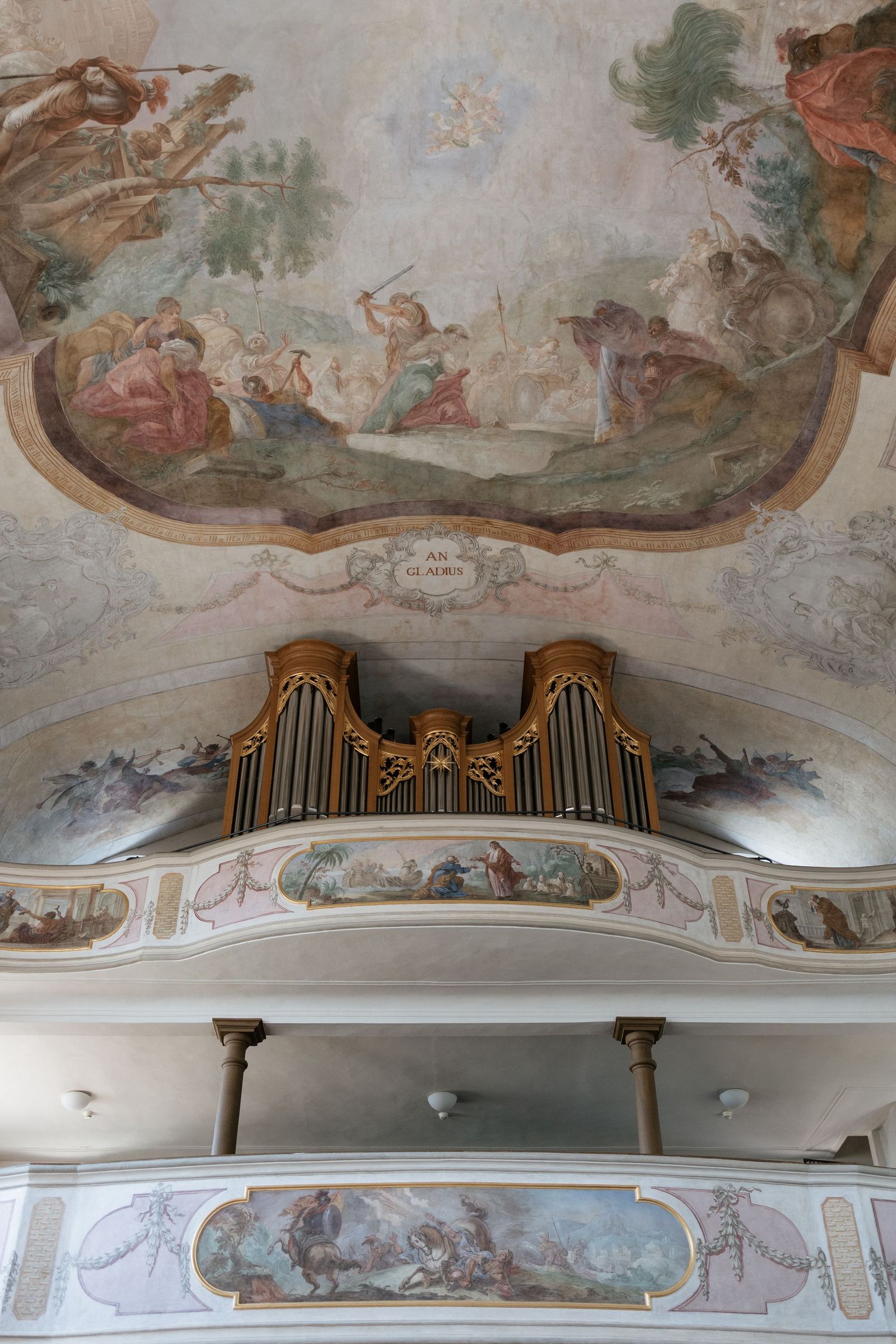 Baroque church interior featuring ornate ceiling frescoes, gilded pipe organ, and decorative painted balcony with religious scenes.