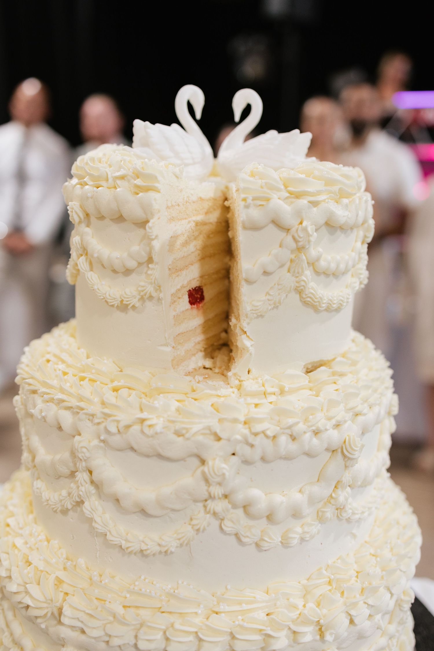Elegant three-tier white wedding cake with swan toppers, intricate frosting details, and a slice cut revealing layers inside.