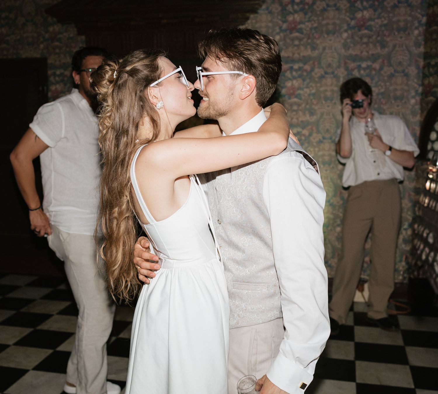 Couple sharing a kiss on the dance floor at a wedding reception, dressed in white and beige attire.