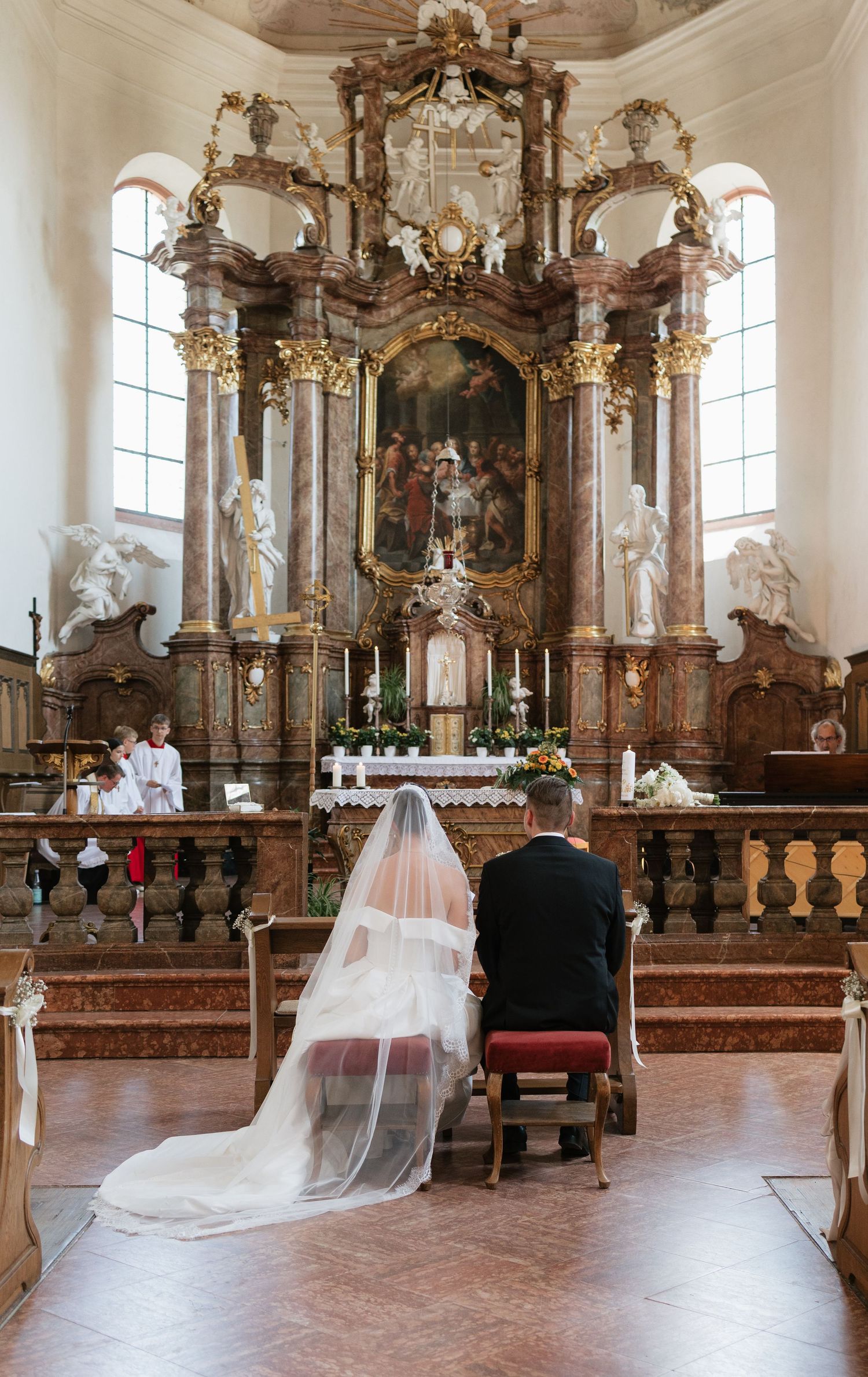 A bride and groom kneel before an ornate Baroque church altar during their wedding ceremony.