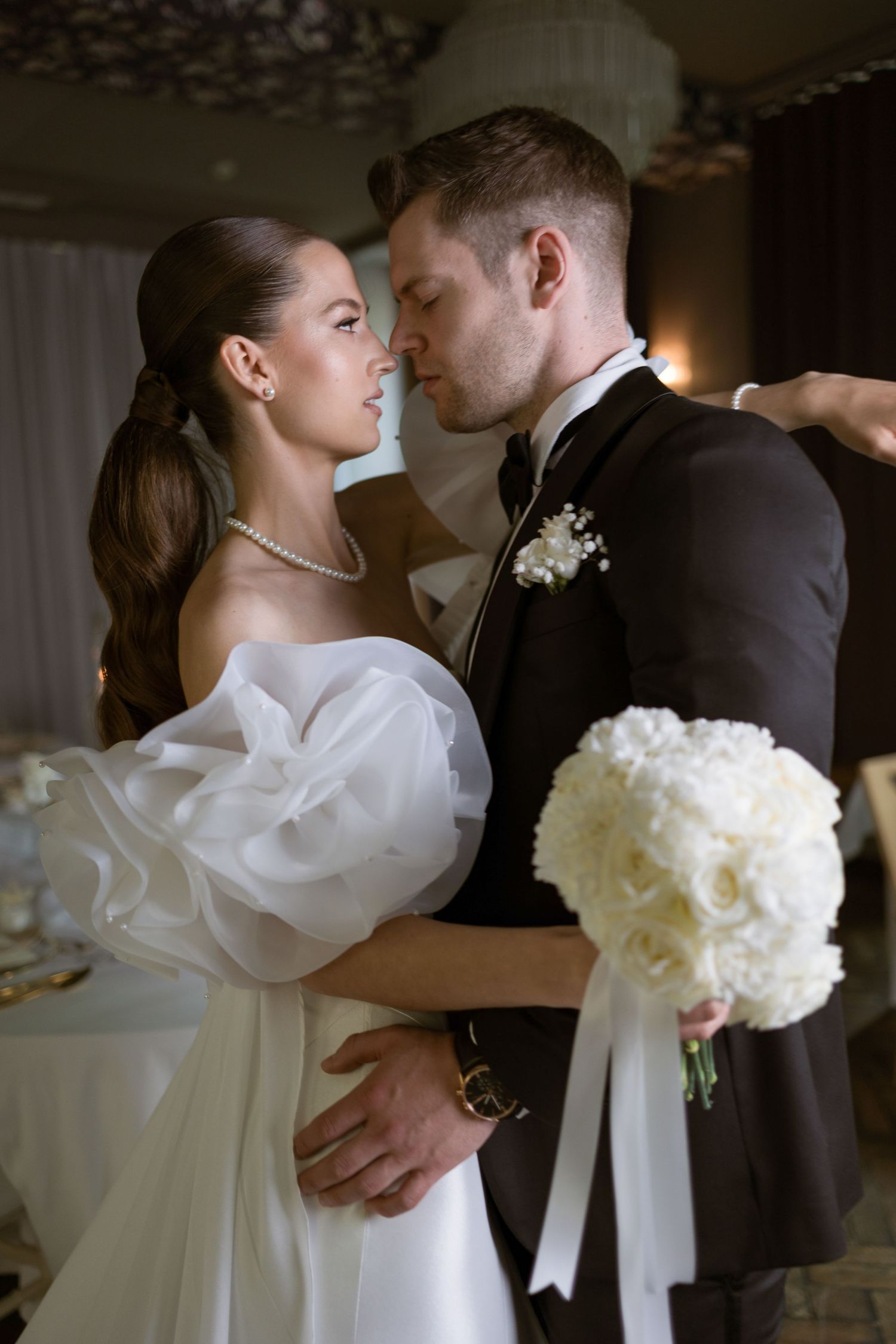 Bride in white gown holding bouquet gazes at groom in black tuxedo during romantic wedding reception.